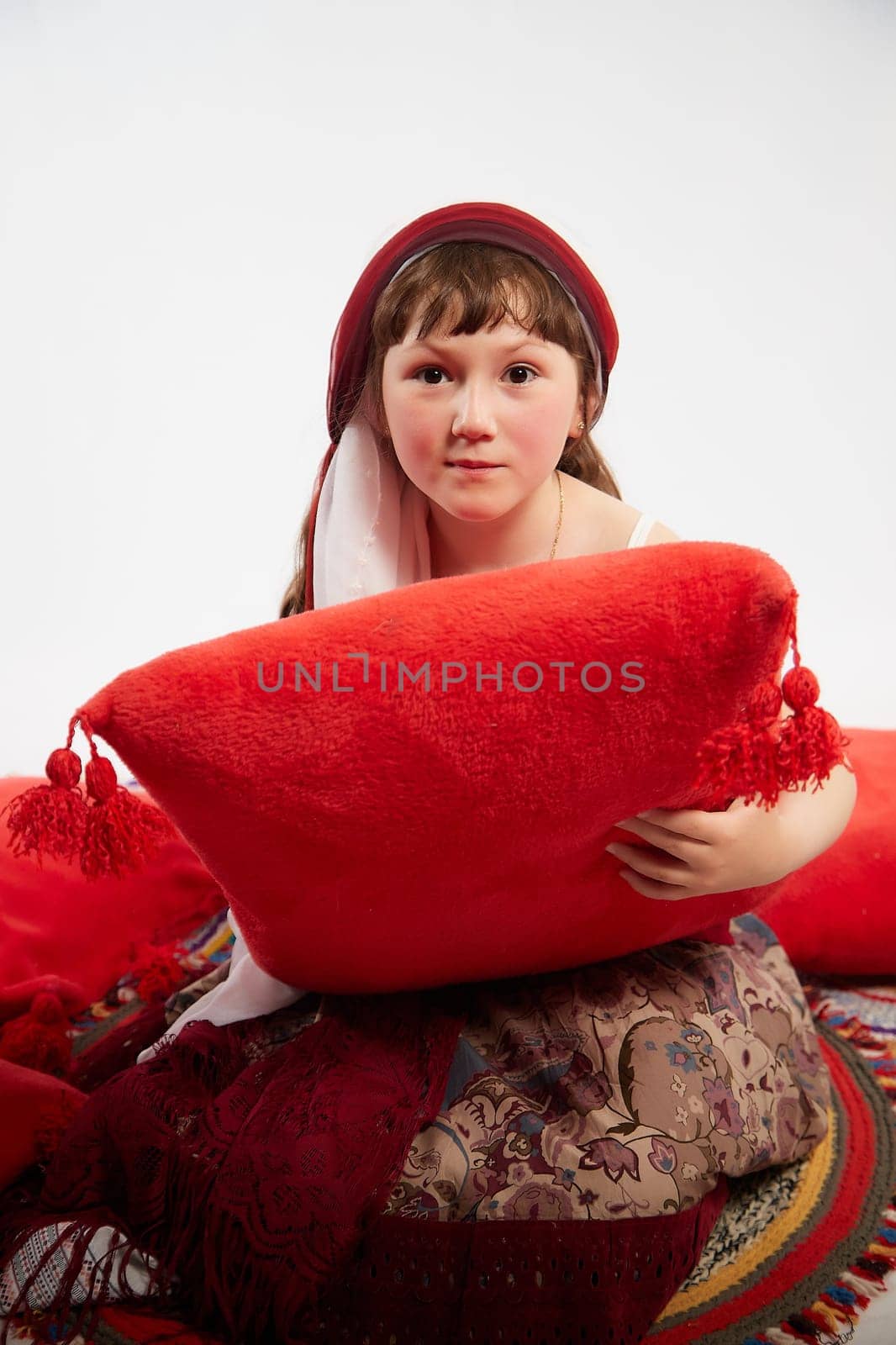 Portrait of Little girl in a stylized Tatar national costume having rest with a red pillow on a white background in the studio. Photo shoot of funny young teenager who is not professional model by keleny