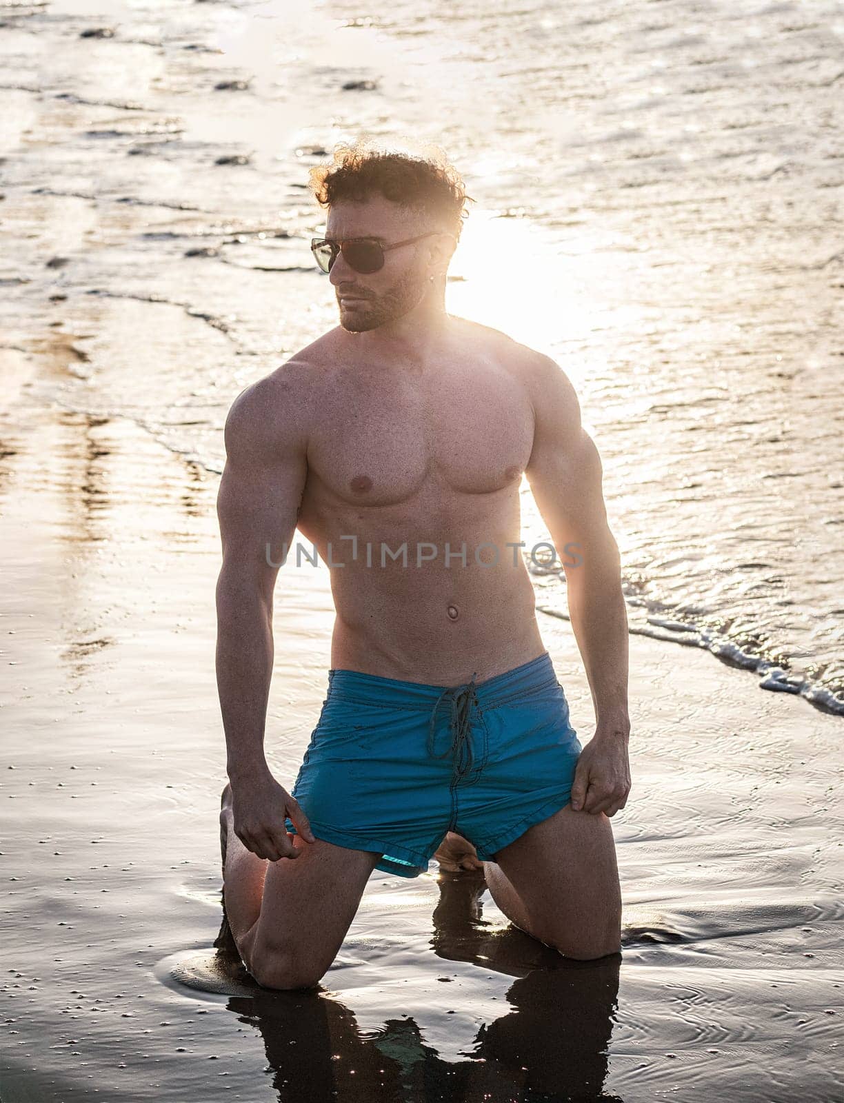 Attractive and fit young bodybuilder in bathing suit kneeling on the beach - Muscular and fit young bodybuilder in the sea