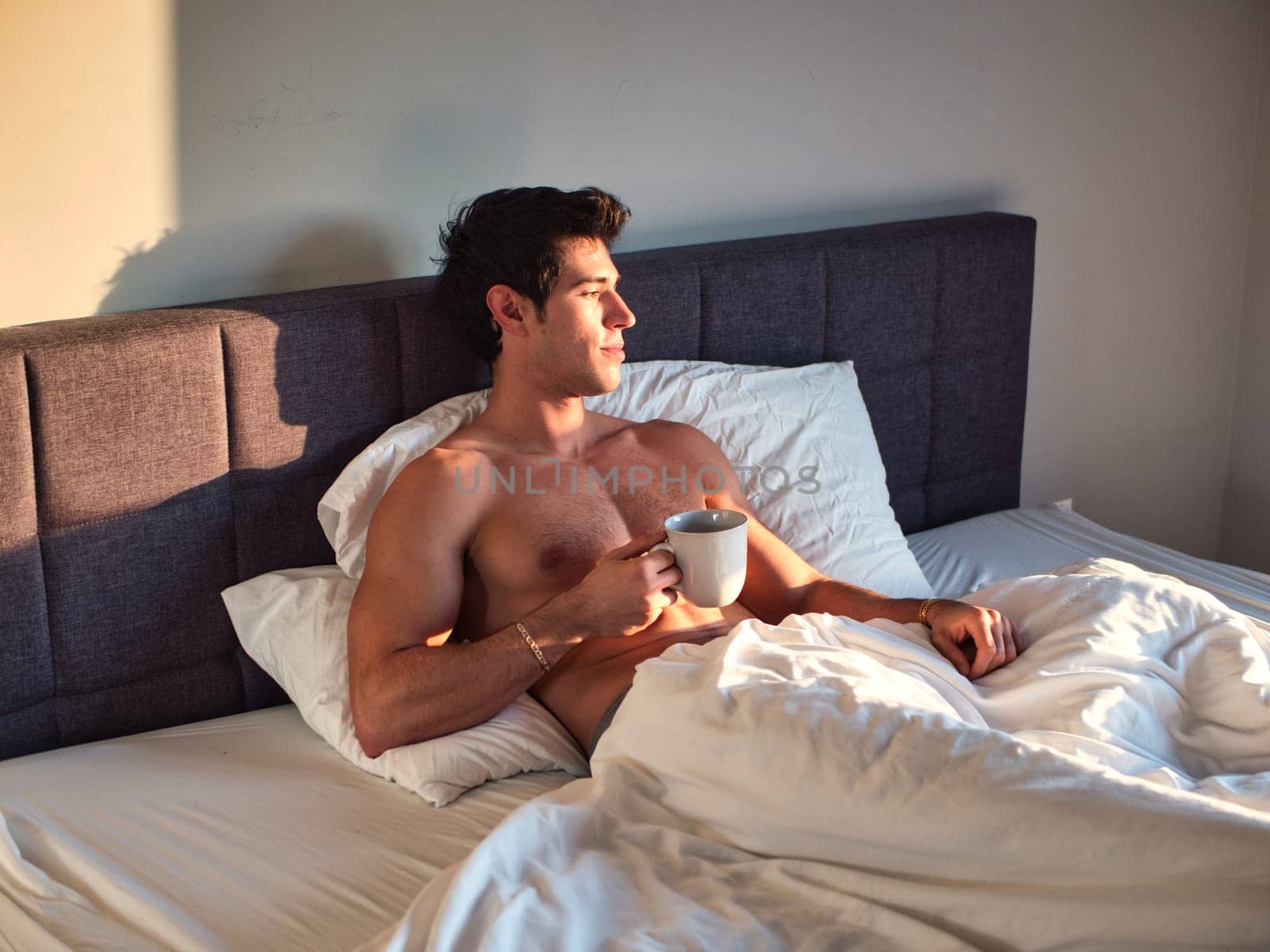 Photo of a shirtless man enjoying a peaceful morning in bed with a cup of coffee by artofphoto
