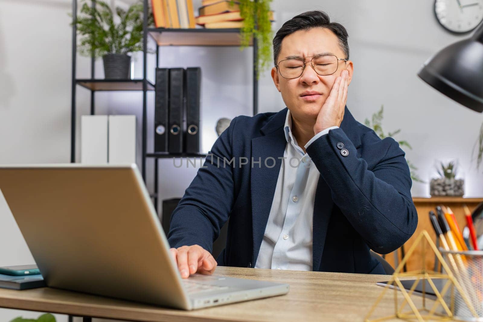 Dental problems. Asian businessman working at office touching cheek, closing eyes with expression of terrible suffer from painful toothache, sensitive teeth, cavities. Male Chinese in formal suit