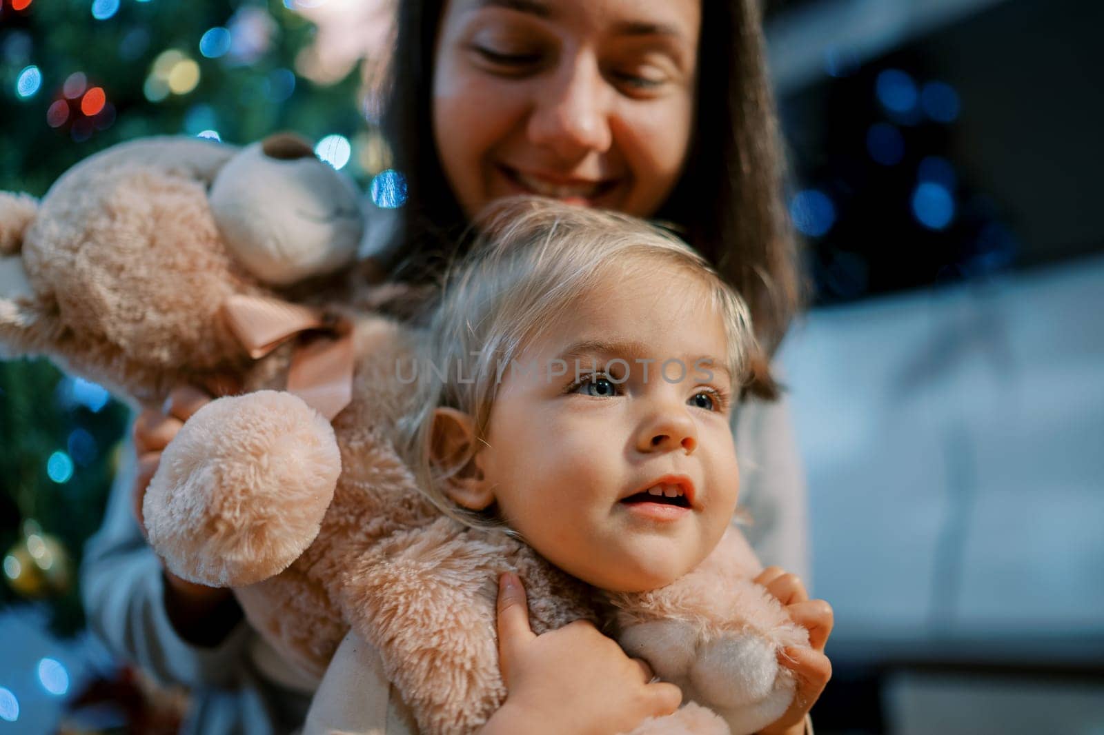 Mom put a teddy bear on the neck of a little girl near the Christmas tree. High quality photo