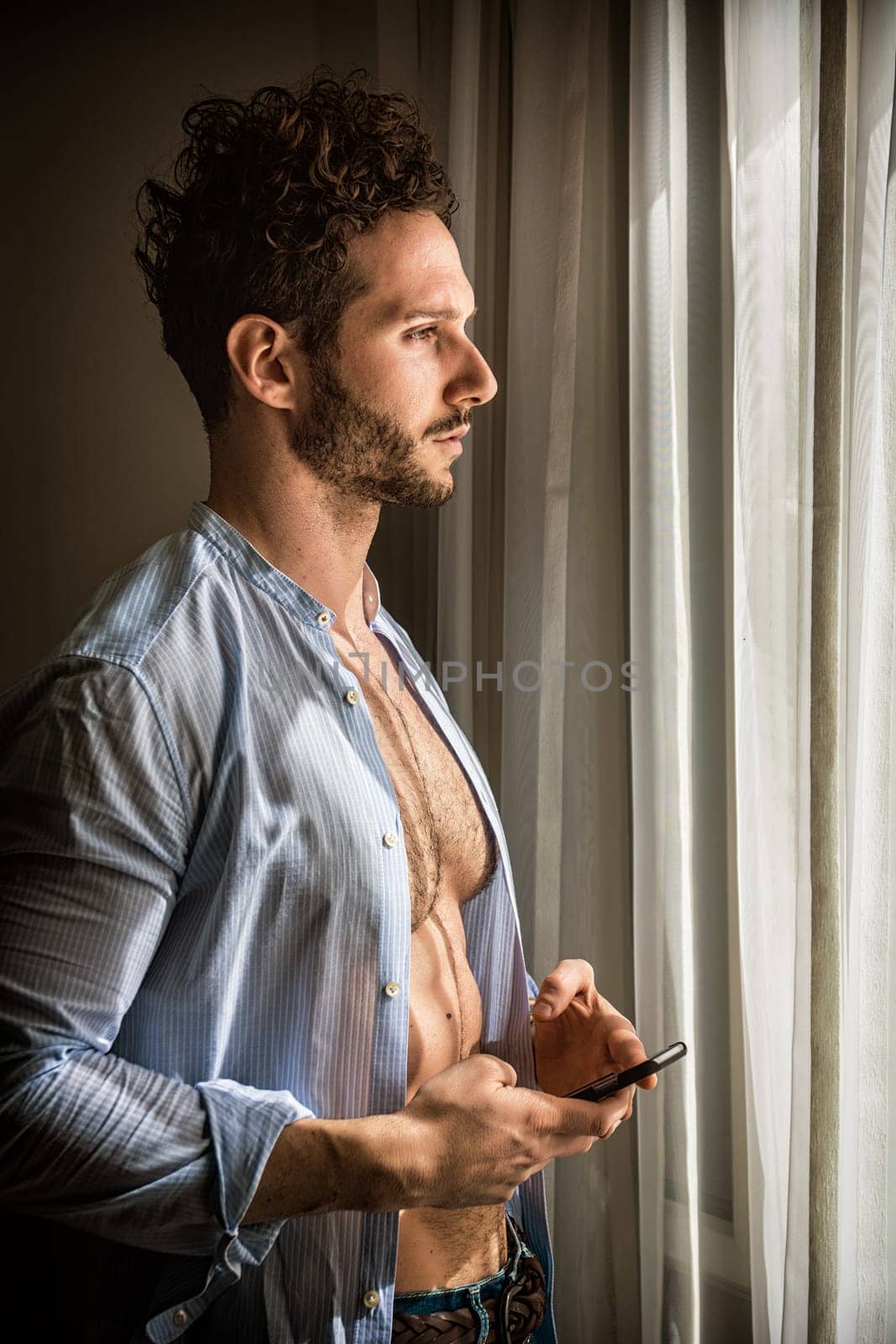A man standing in front of a window holding a cell phone. A Captivating View: A Muscular Man Captivated by His Cell Phone