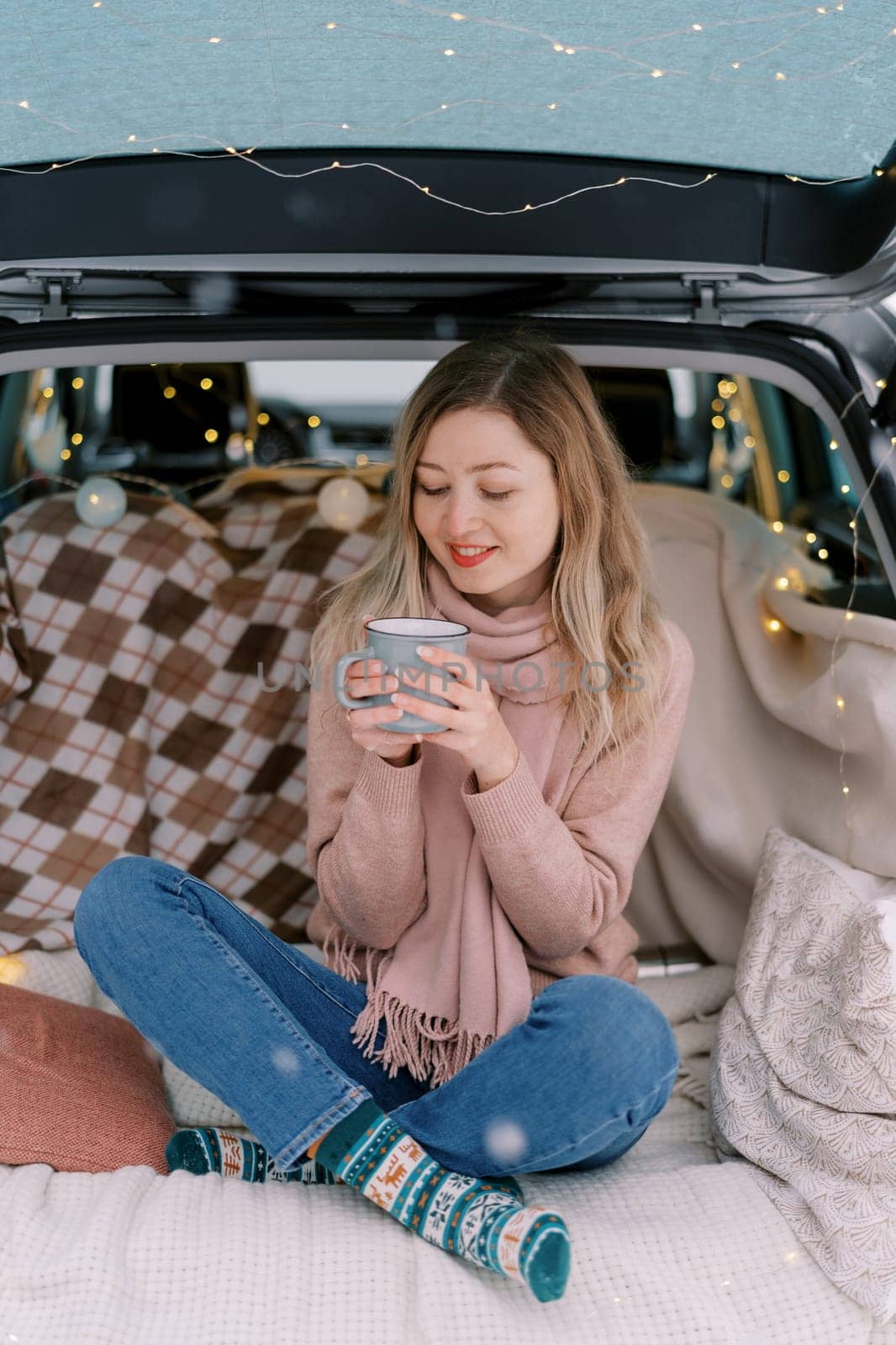 Smiling girl sits cross-legged on a blanket in the trunk of a car and looks at a cup of coffee in her hands. High quality photo