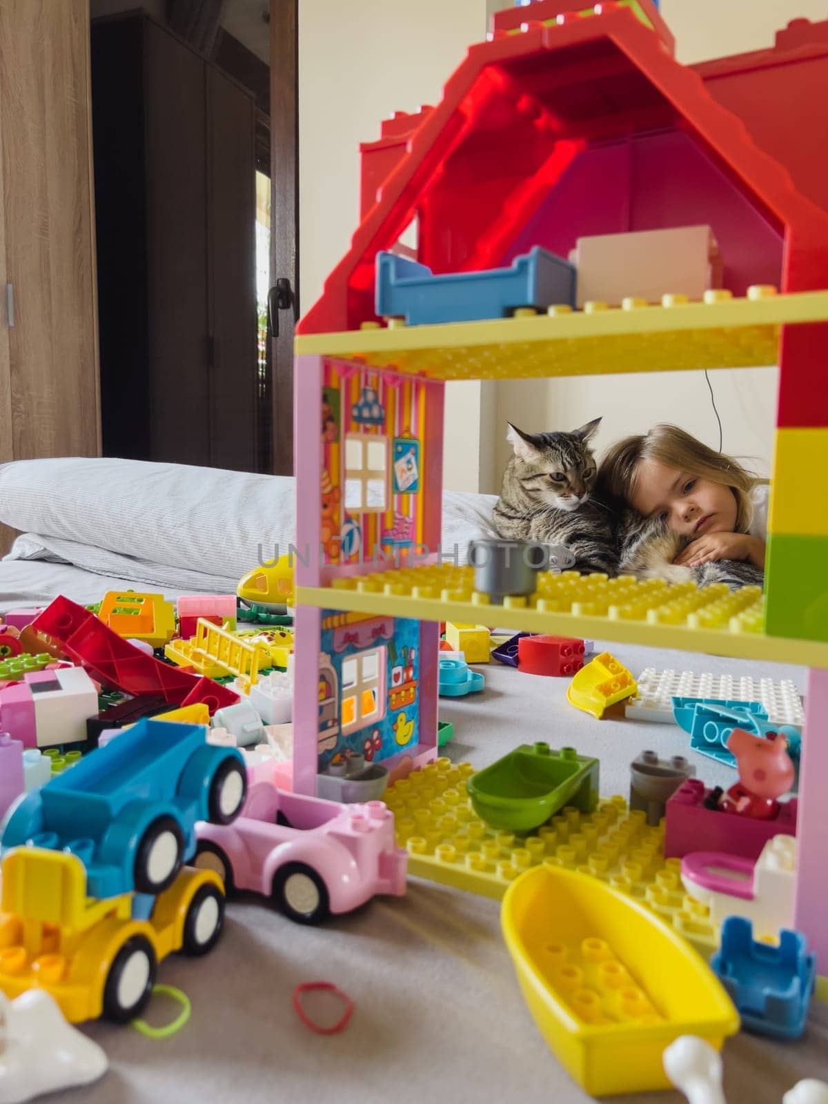 Little girl hugs a tabby cat lying on a bed near a colorful lego house. High quality photo