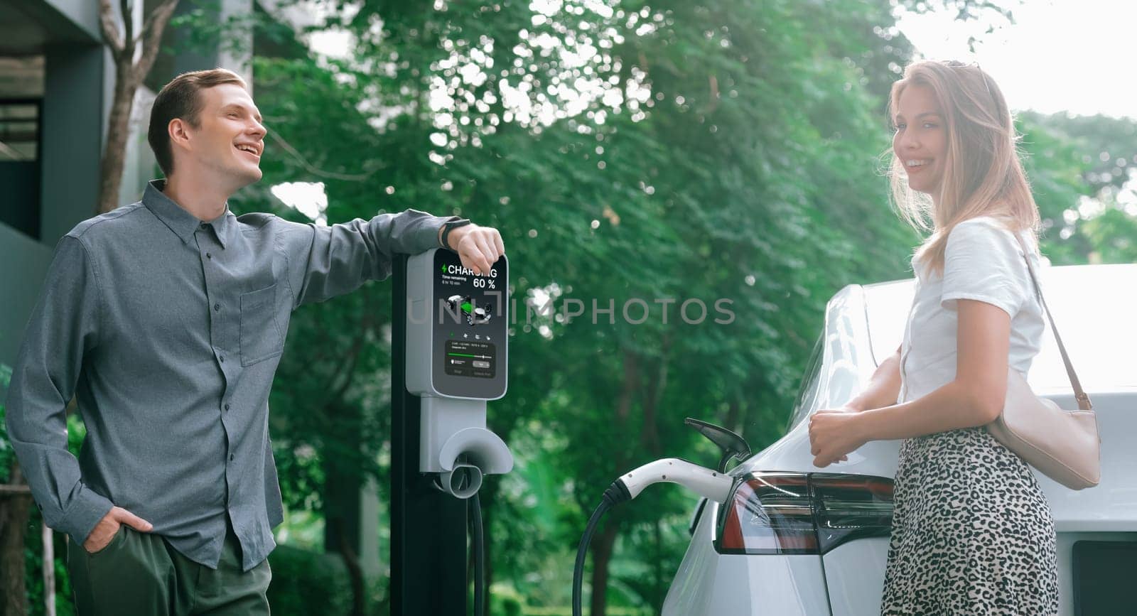 Young couple travel with EV electric car charging in green sustainable city outdoor garden in summer shows urban sustainability lifestyle by green clean rechargeable energy of electric vehicle innards