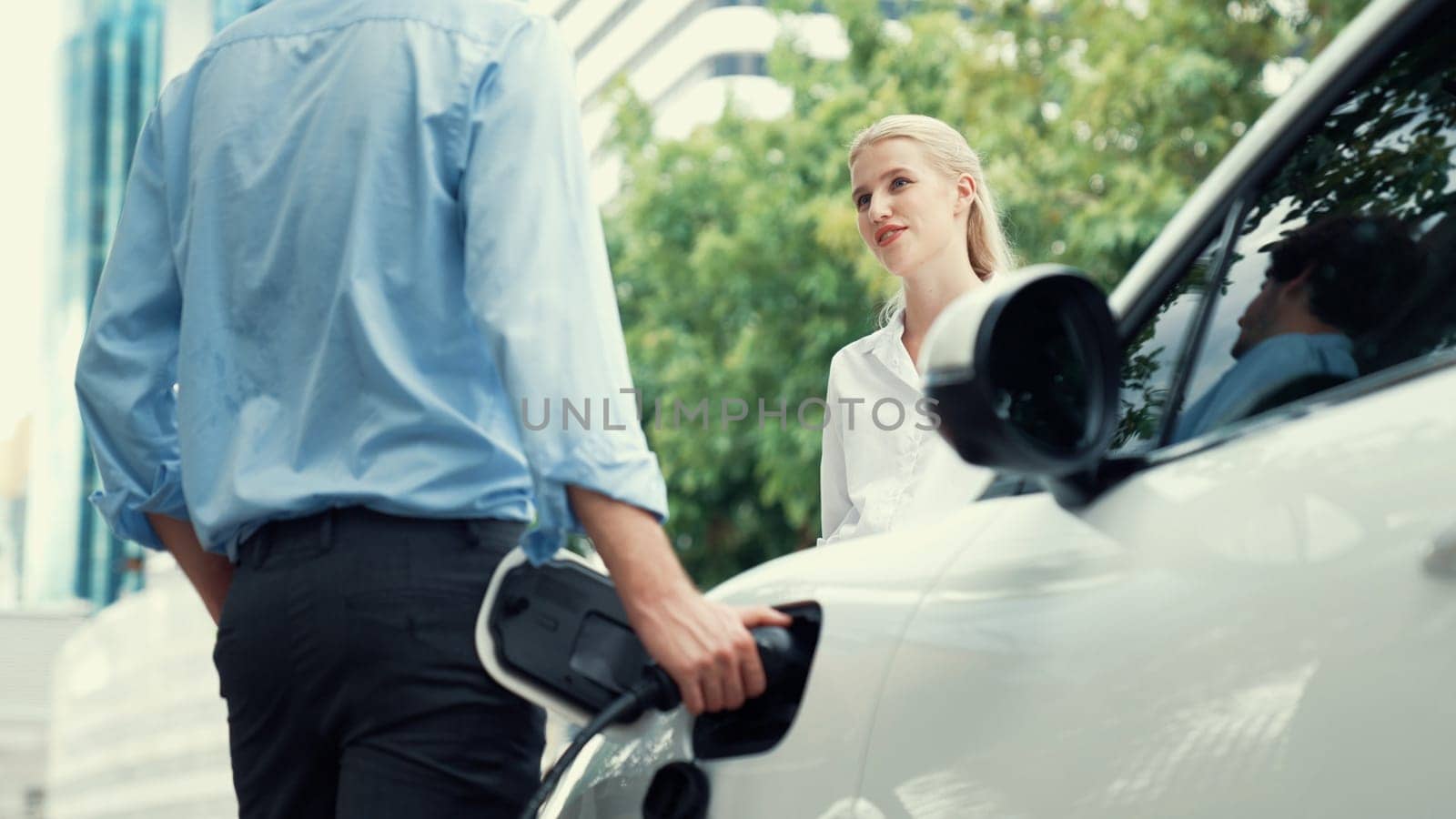 Progressive businessman and businesswoman install charger plug from charging station to electric car before driving around city center. Eco friendly rechargeable car powered by clean energy.