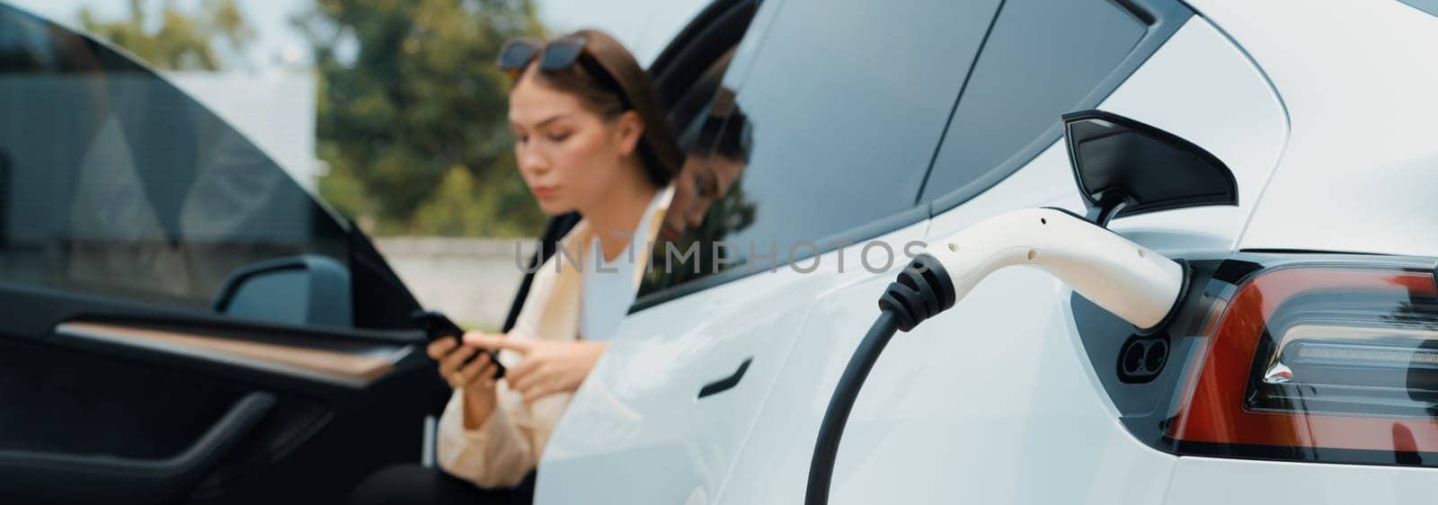 Young woman use smartphone to pay for electricity at public EV car charging station green city park. Modern environmental and sustainable urban lifestyle with EV vehicle. Panorama Expedient
