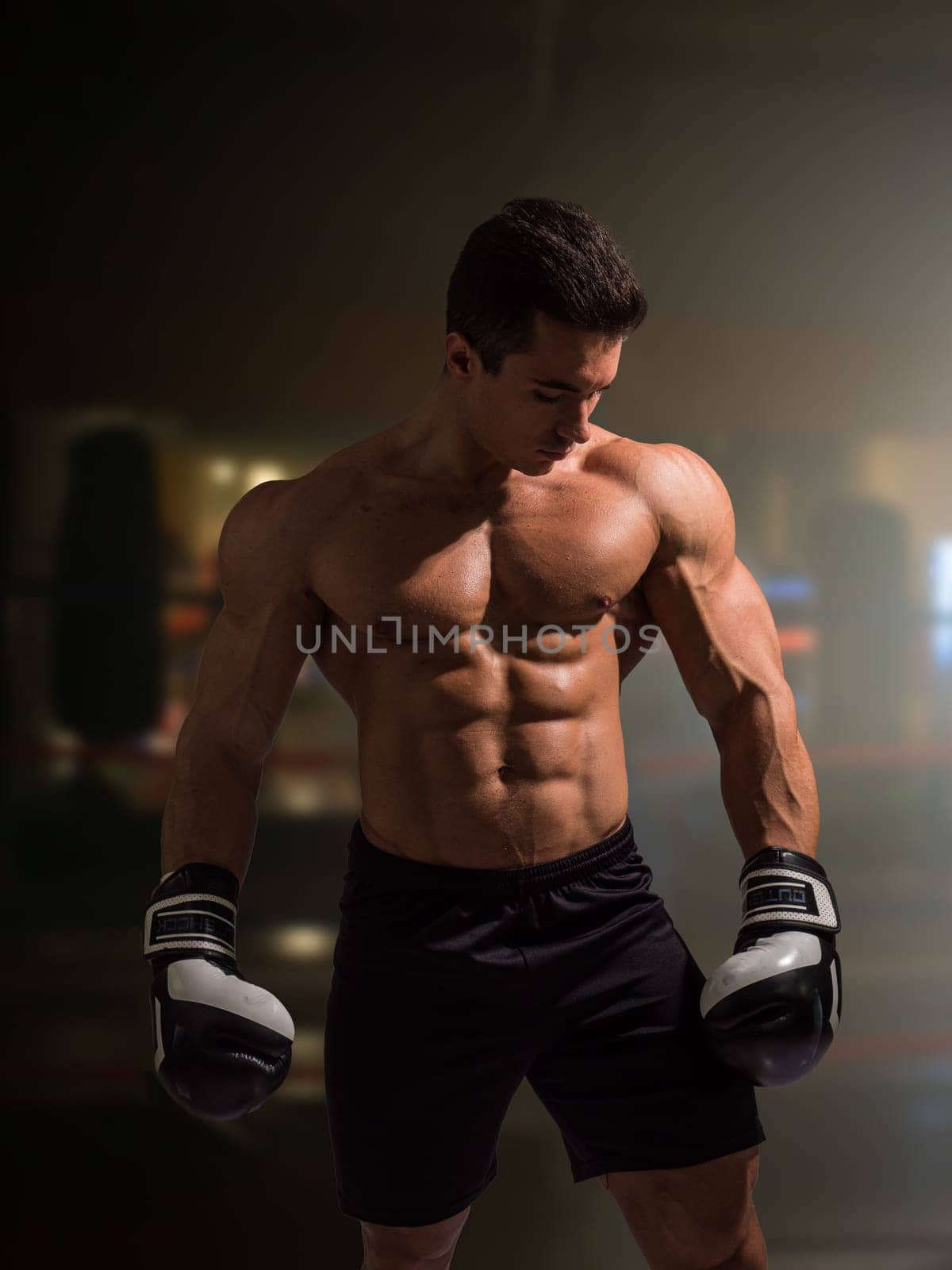 A muscular young man in gym with no shirt and boxing gloves