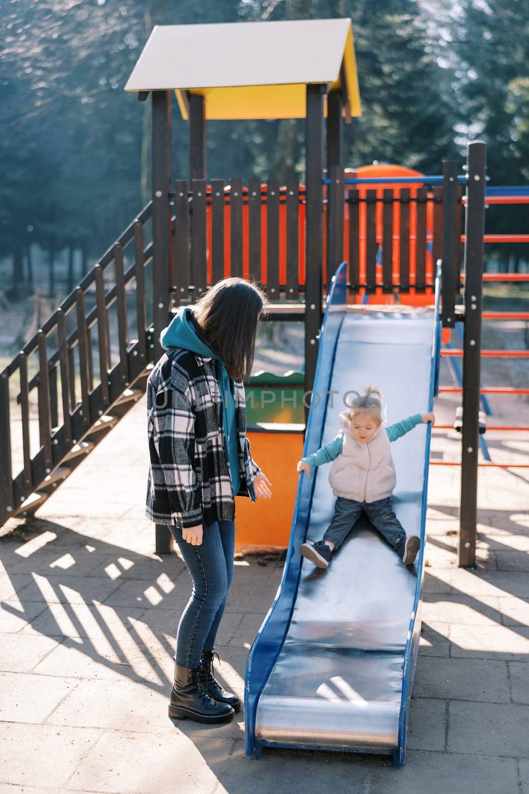 Little girl goes down the slide on the playground next to her standing mom. High quality photo