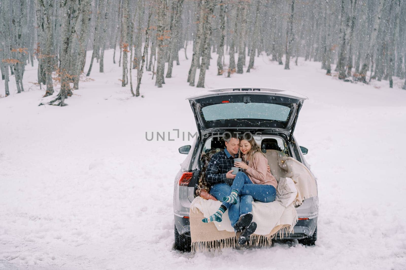 Couple with cups of coffee sits hugging in the trunk of a car on blankets under snowfall in the forest. High quality photo