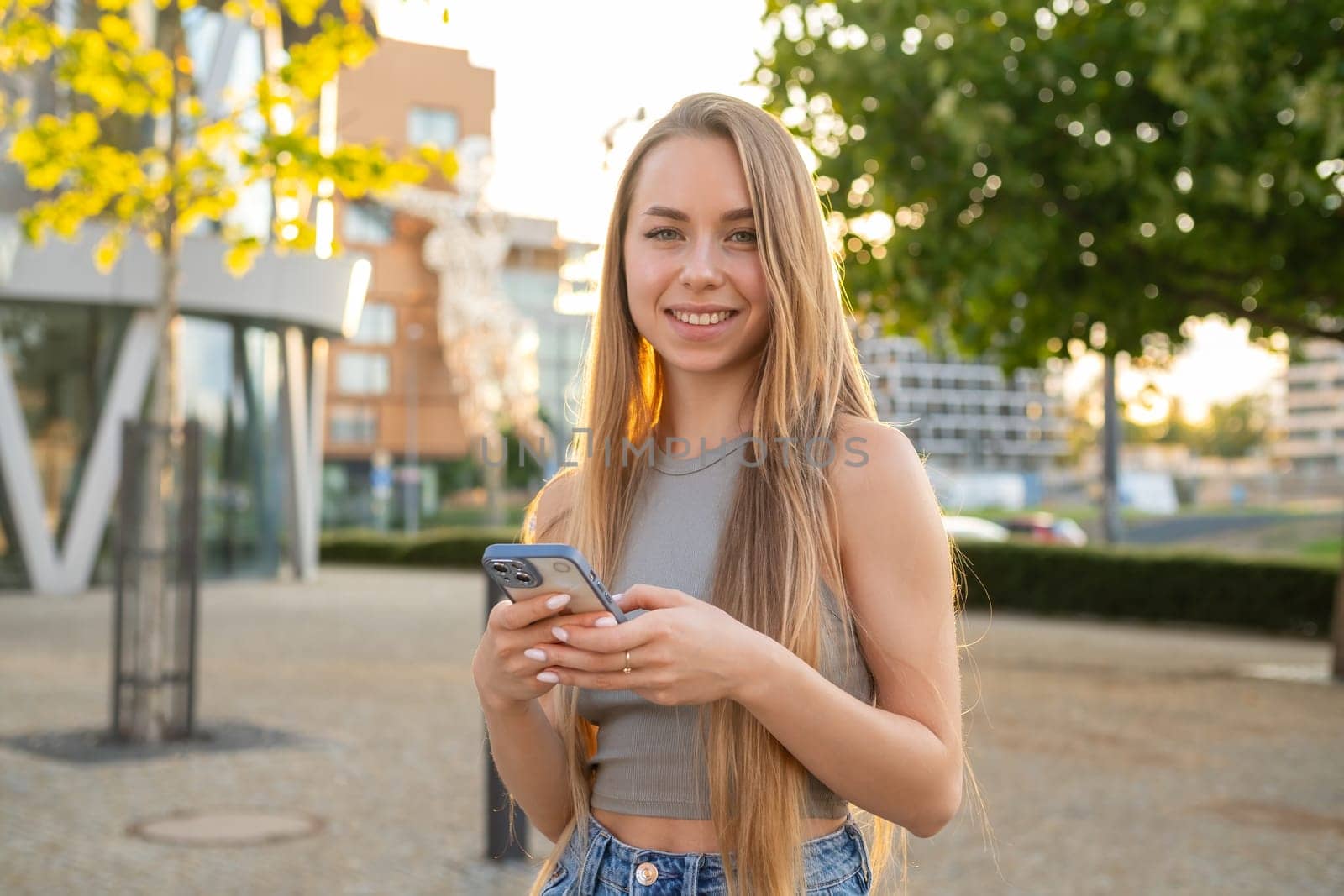 Portrait of young blonde hair woman with beautiful smile standing in the street and uses mobile phone.