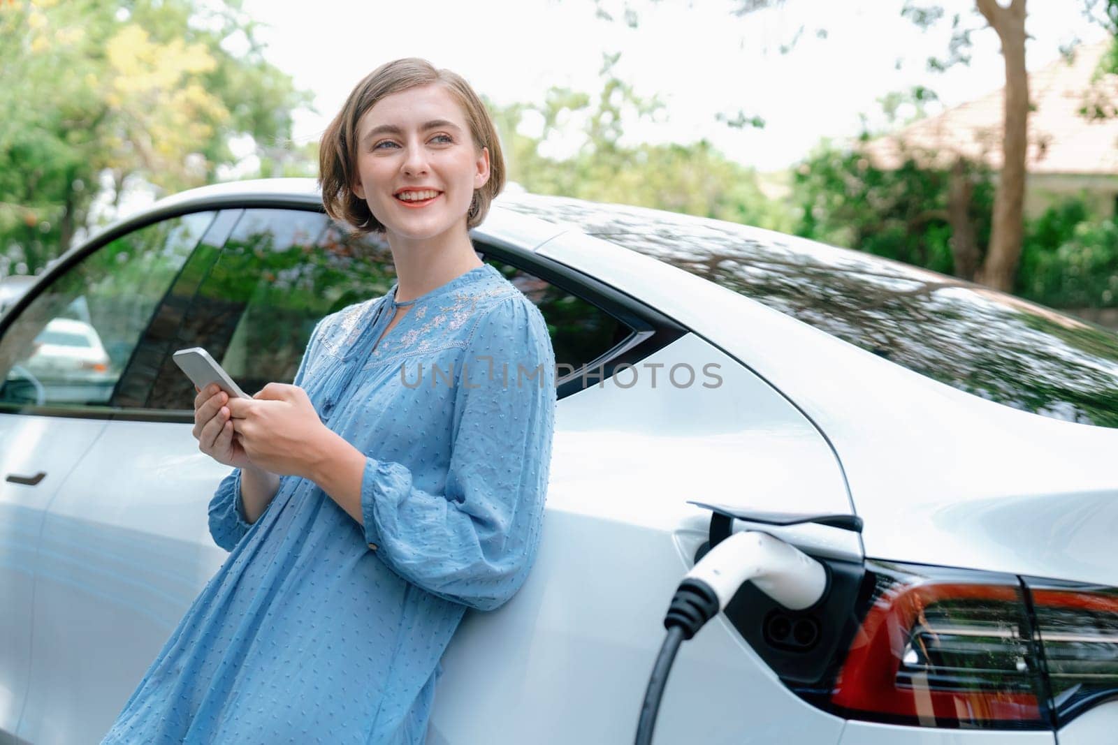 Holiday road trip vacation by the green countryside nature with beautiful young woman checking battery status from smartphone while recharging electric vehicle.Eco-friendly travel wit EV car.Perpetual