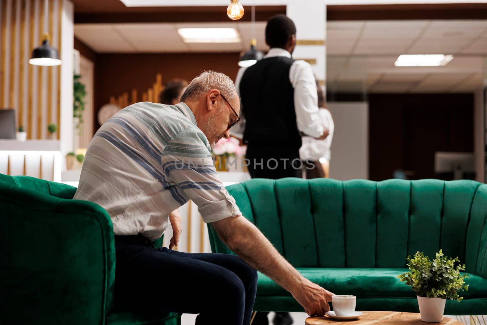 At elegant lounge area for check-in, elderly caucasian man sits on sofa reaching for cup of coffee on table. Senior guy in hotel lobby, waiting on the couch for the booking procedure.