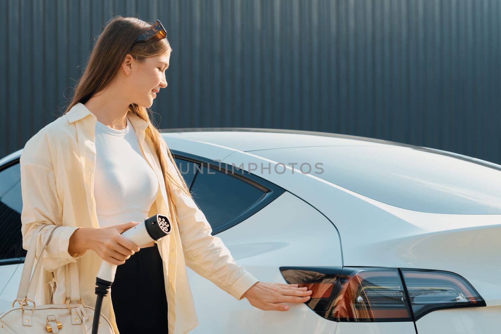 Young woman holding shopping bag recharge EV car battery from charging station at parking lot. Modern woman go shopping by environmental friendly electric vehicle in urban travel lifestyle. Expedient