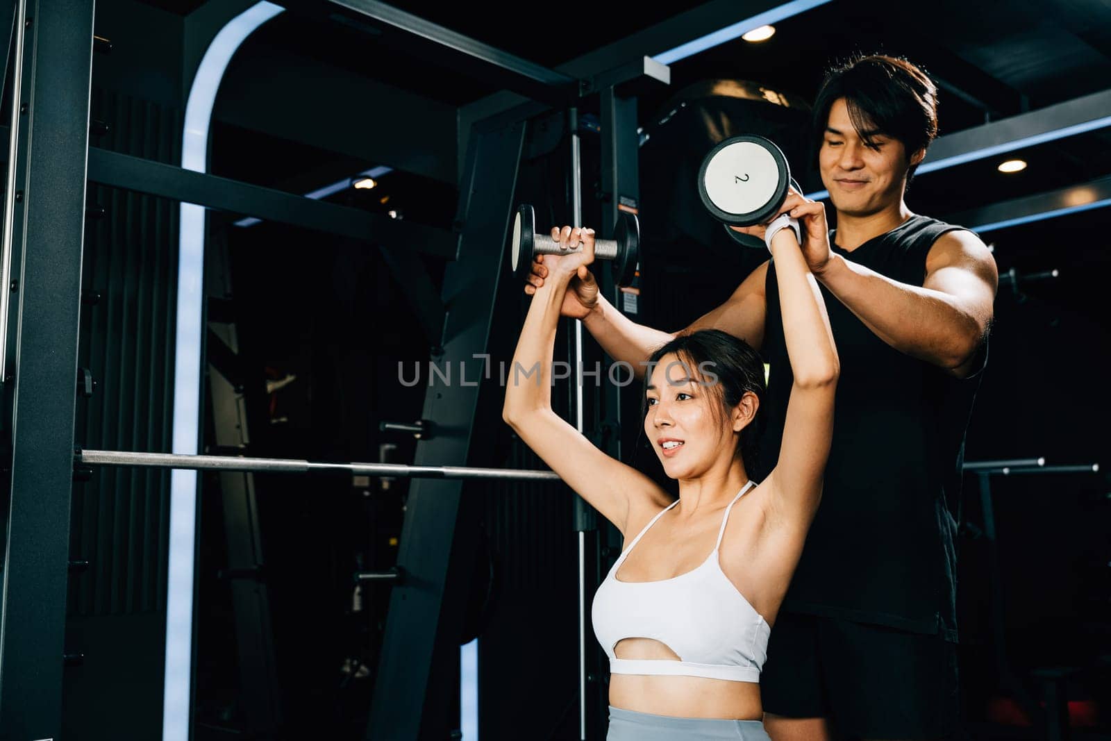 Fit Asian woman with a muscular and toned arm lifting weights in the gym, motivated by her personal trainer's guidance and support and help, technique of exercise in gym dark gym background