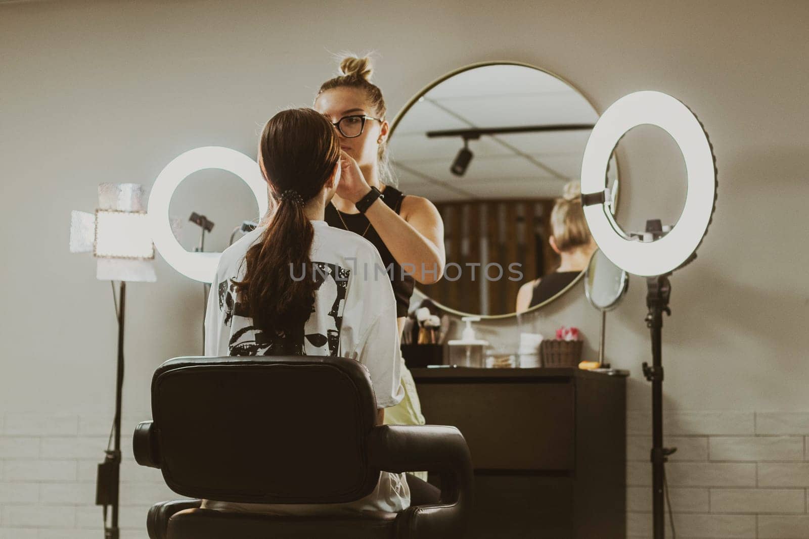 One young beautiful makeup artist does makeup to a girl sitting in a chair from the back early in the morning in a beauty salon, close-up side view.Step by step.