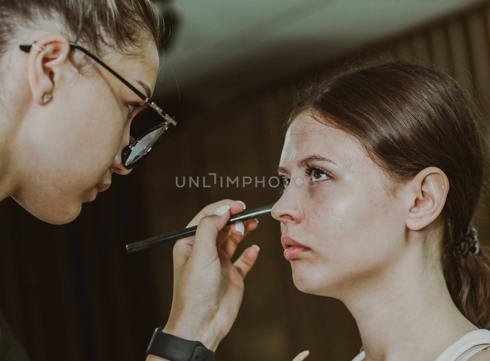 One young handsome Caucasian makeup artist applies eye shadow to the lower eyelid of a girl sitting in a chair early in the morning in a beauty salon, close-up side view. Step by step.