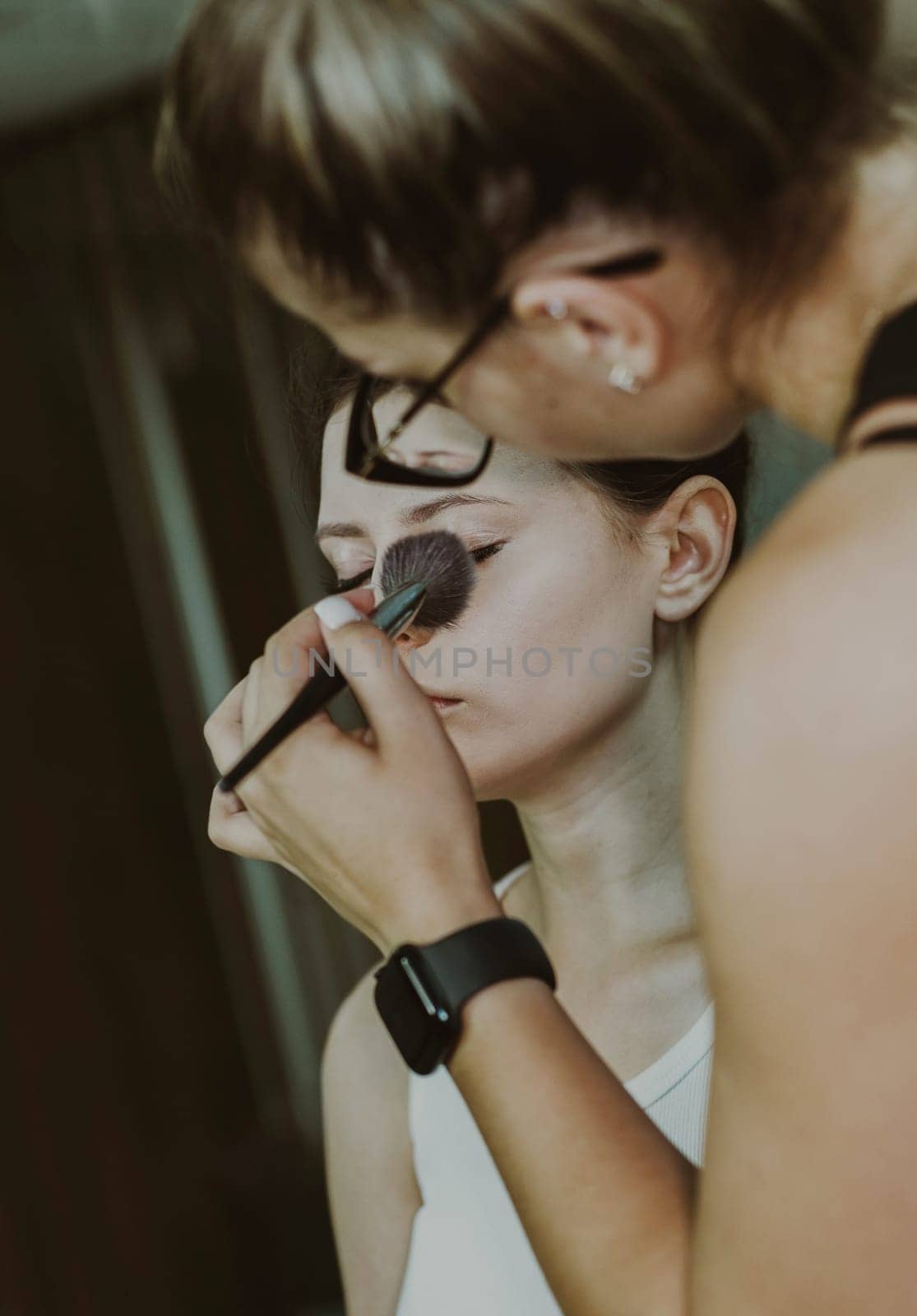 One young handsome Caucasian makeup artist applies foundation powder with a fluffy brush to the eyes of a girl sitting in a chair early in the morning in a beauty salon, close-up side view. Step by step.