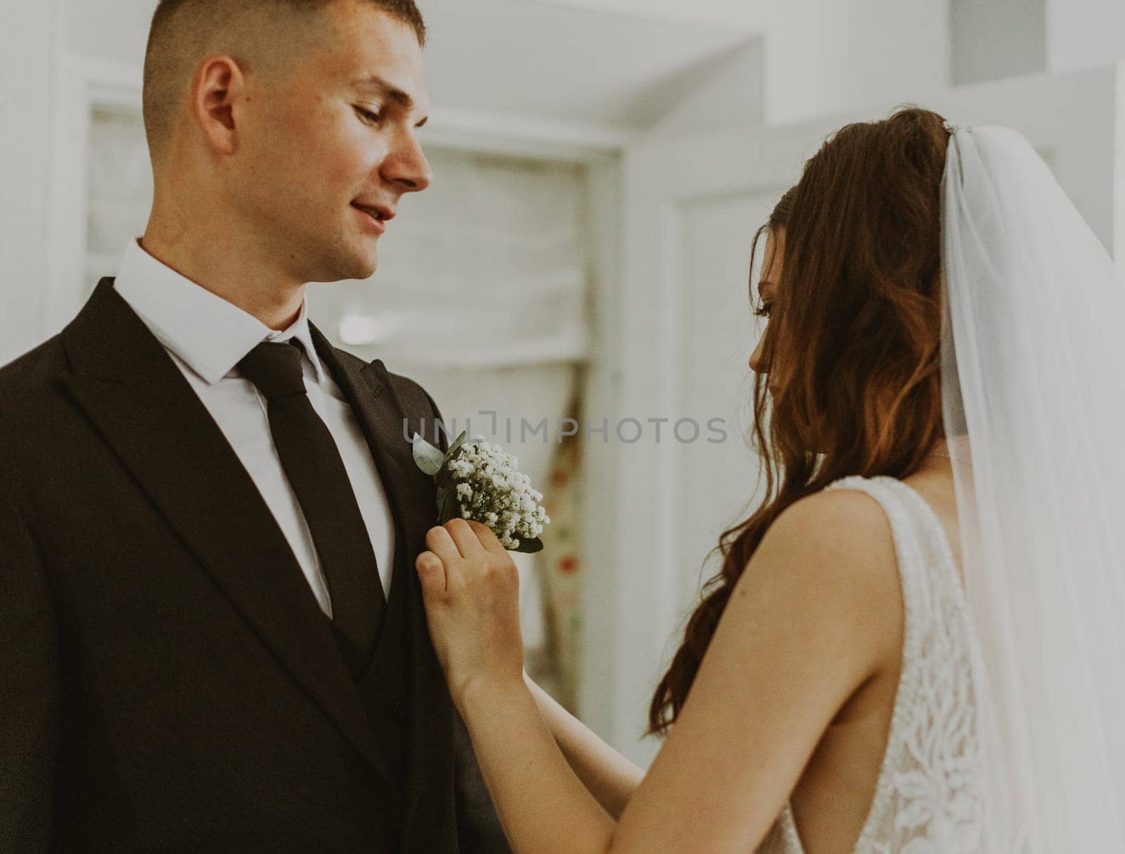 One young handsome Caucasian bride inserts a small bouquet of white boutonnieres into the pocket of the groom's jacket, preparing for the wedding, side view close-up.