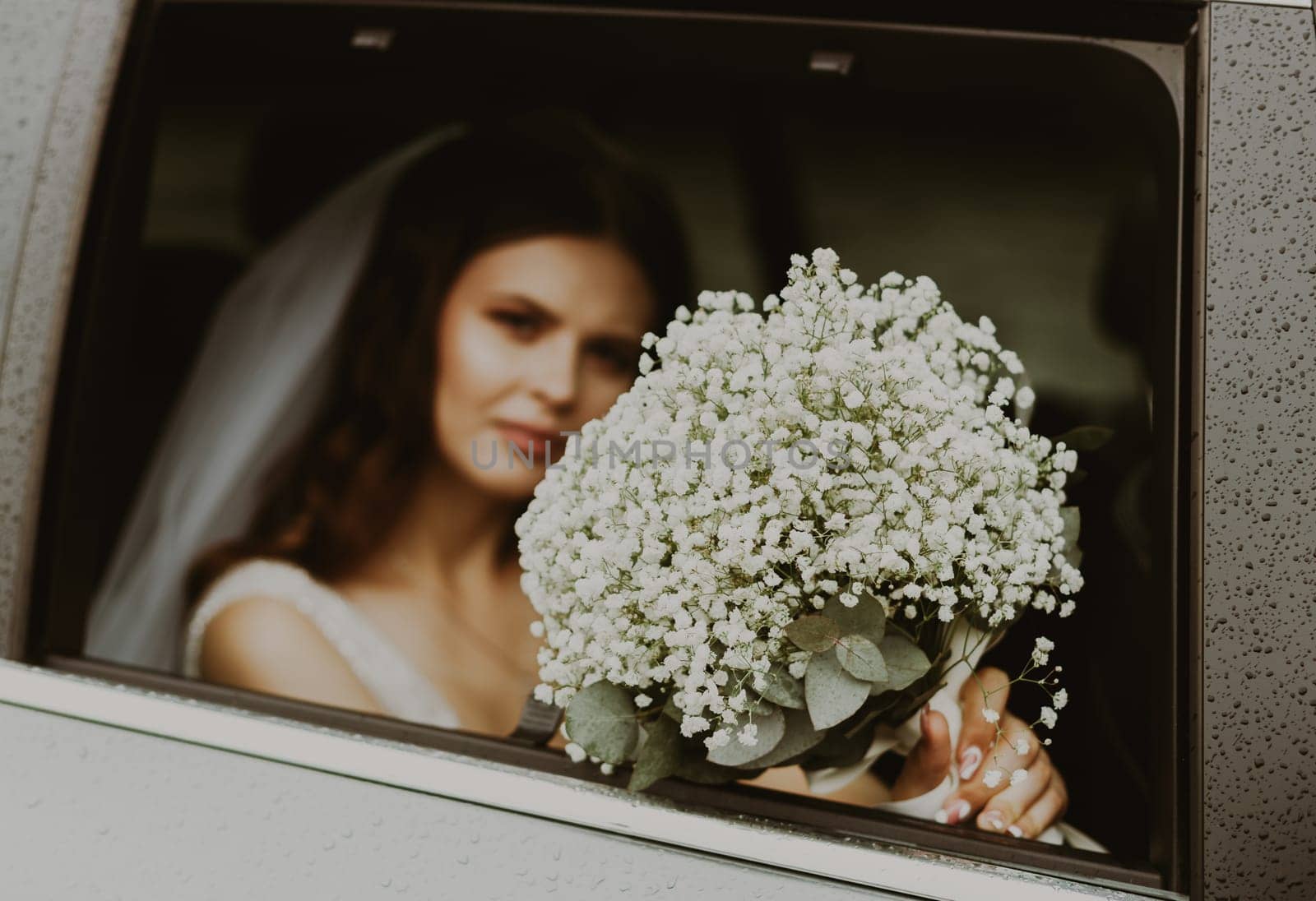 One young beautiful Caucasian bride holding out a large bouquet of white boutonnieres out the car window on a cloudy rainy day, close-up side view with selective focus and depth of field.
