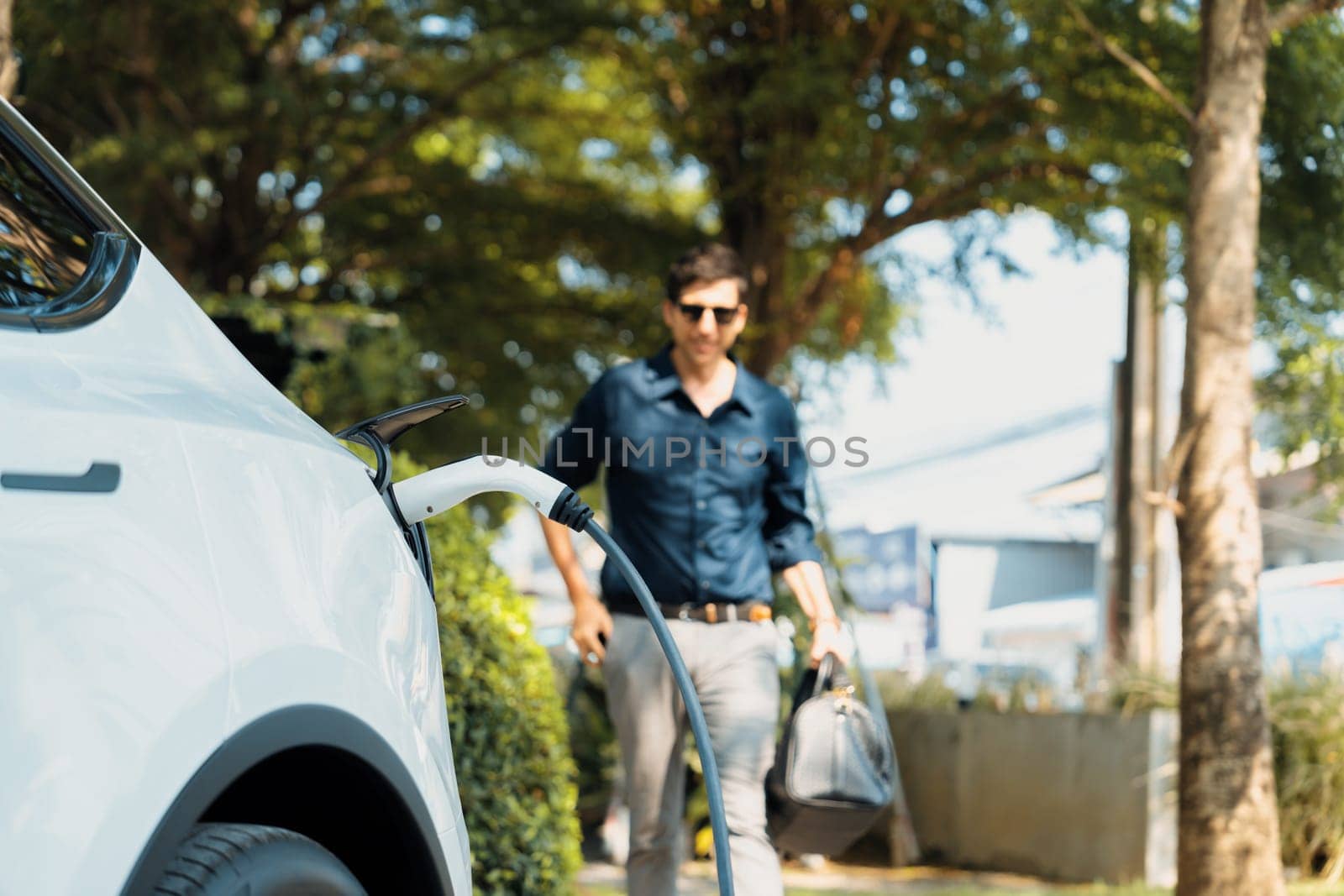 Young man recharge electric car's battery from charging station in outdoor green city park in springtime. Rechargeable EV car for sustainable environmental friendly urban travel lifestyle. Expedient