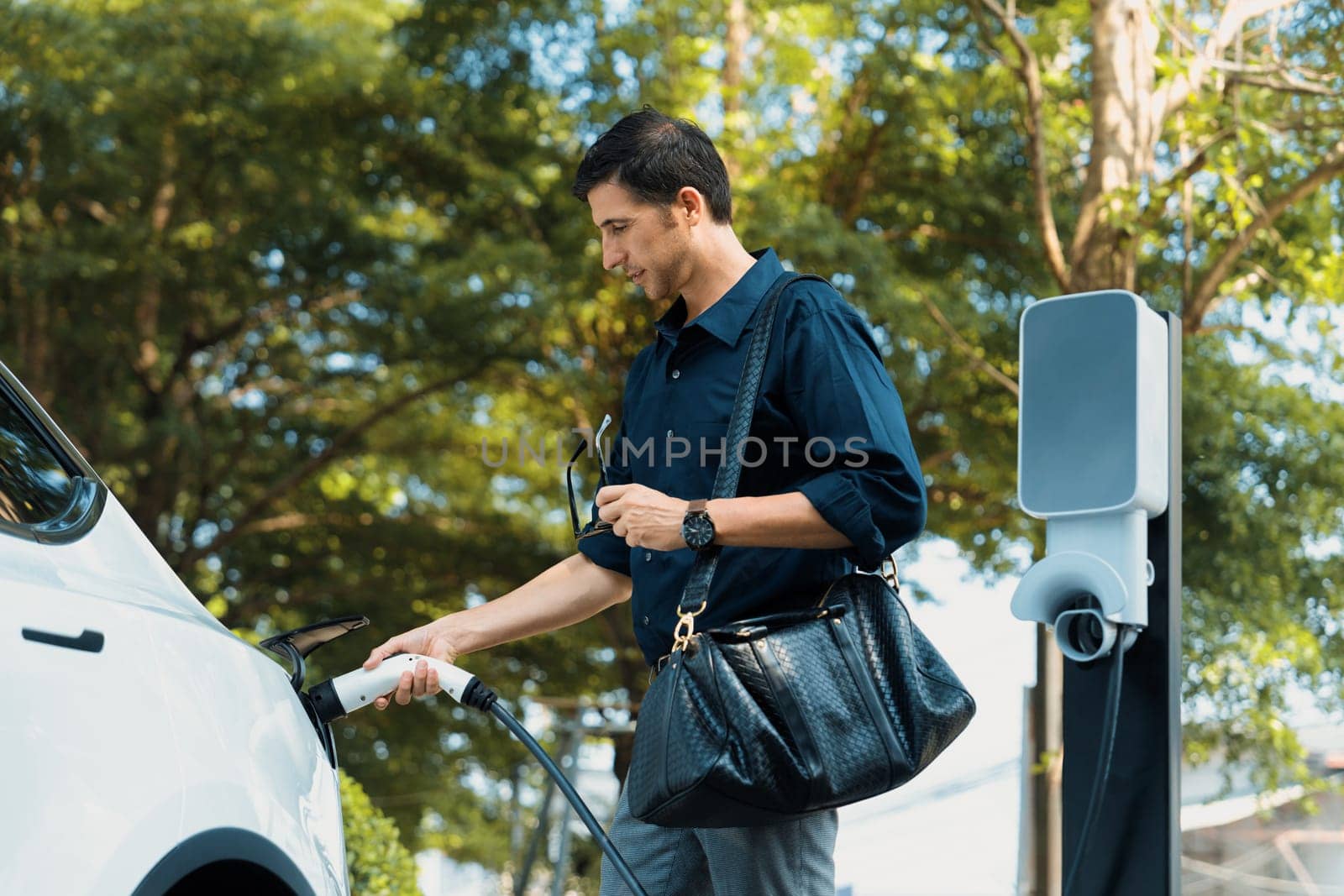 Young man recharge electric car's battery from charging station in outdoor green city park in springtime. Rechargeable EV car for sustainable environmental friendly urban travel lifestyle. Expedient