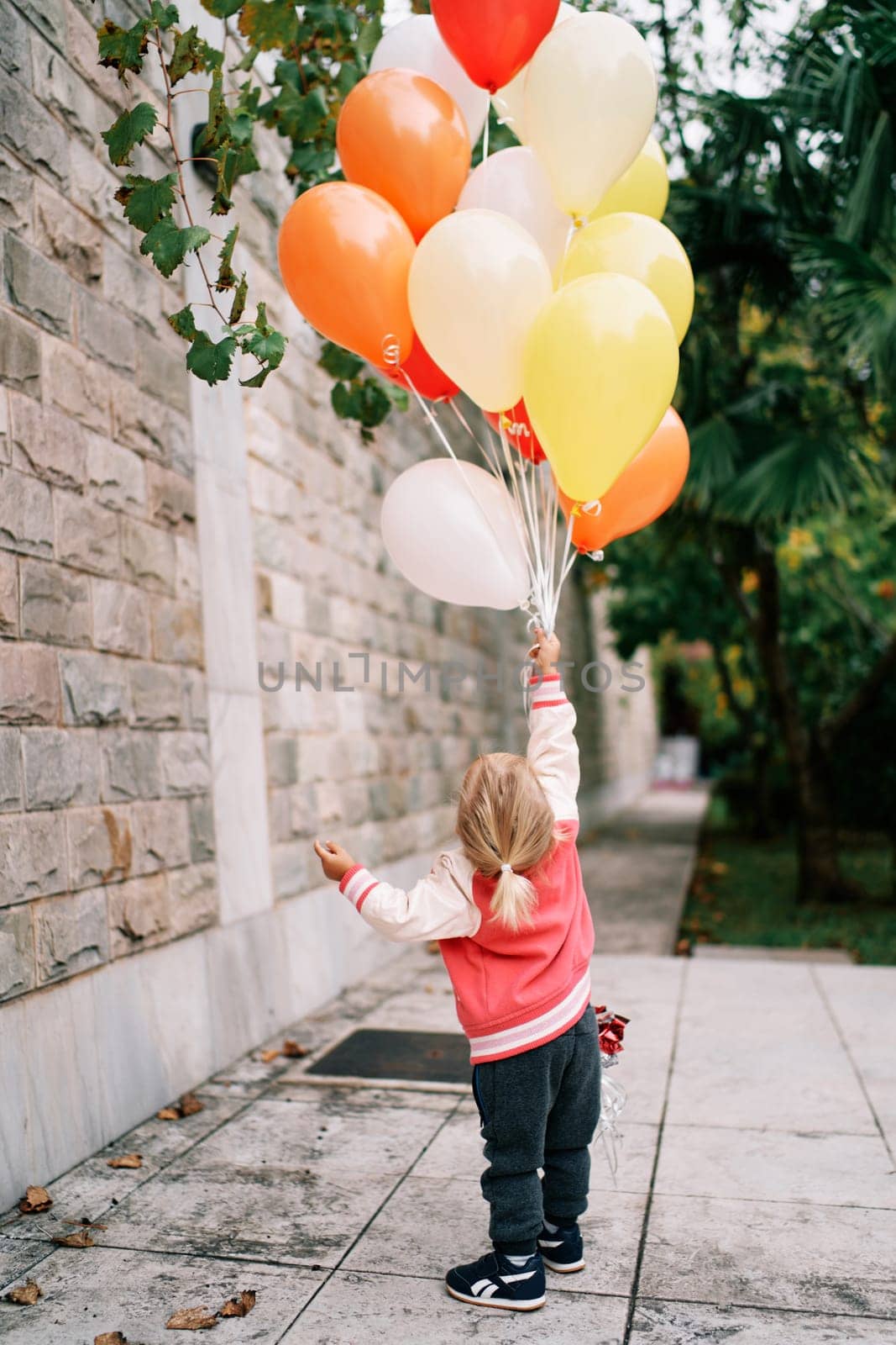 Little girl with a bunch of balloons stands against the wall in the garden and looks up. Back view. High quality photo
