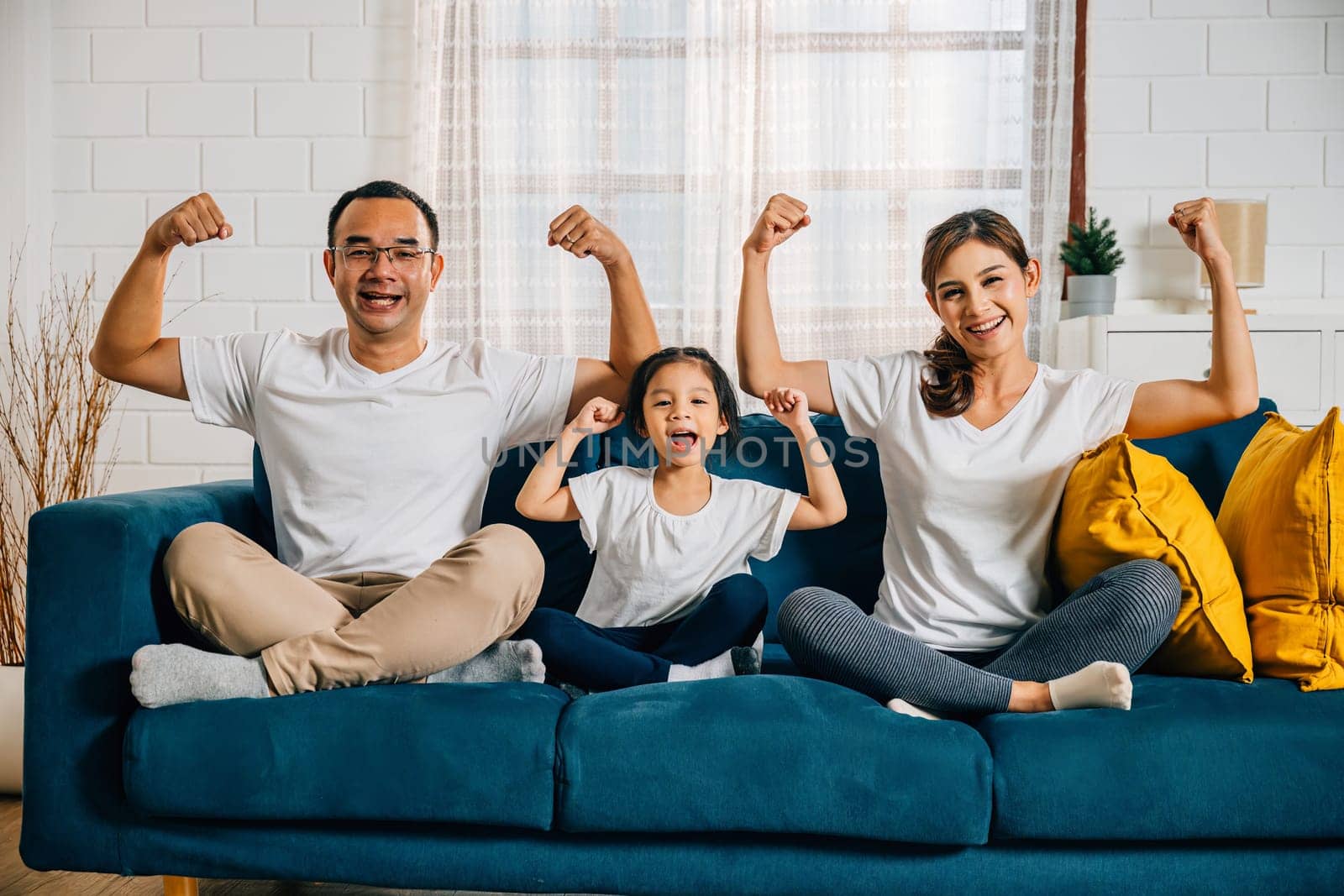 In their cozy living room a family day concept unfolds as young parents their child and daughter highlight their biceps muscles showcasing strength and togetherness with smiles and joy.