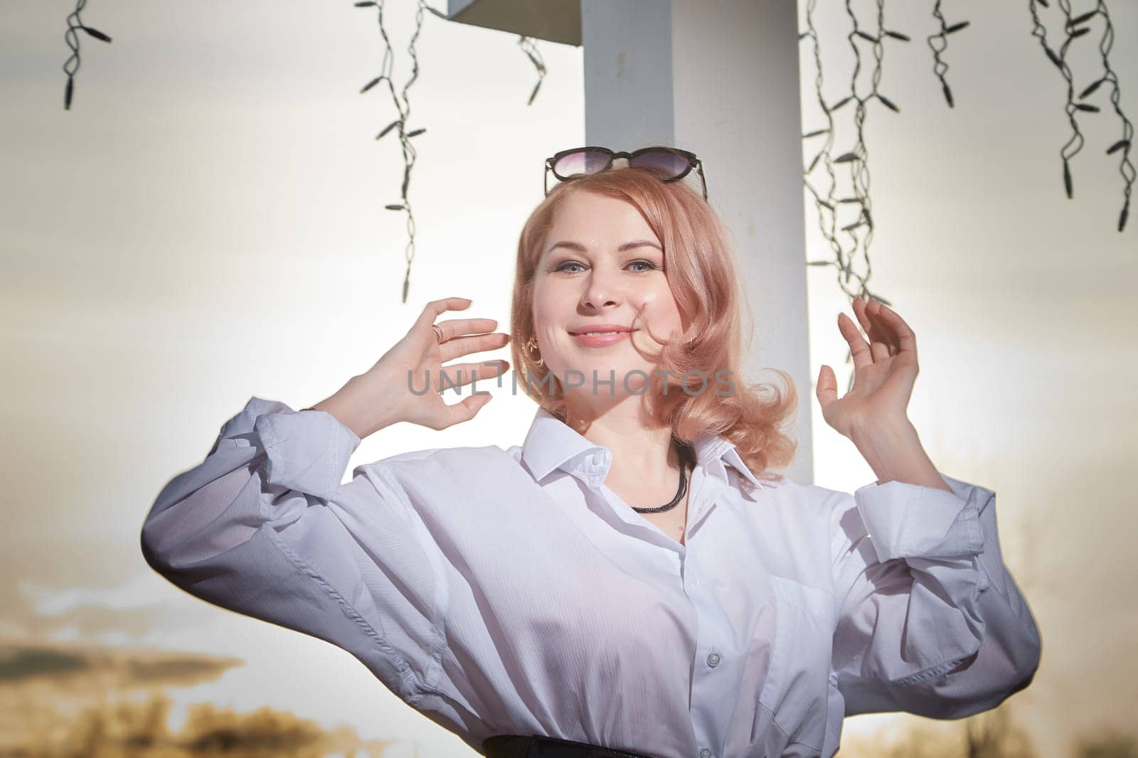 Portrait of Beautiful girl with red hair in white shirt in open wooden pavillion in village or small town. Young slender woman and sky with clouds on background on autumn, spring or summer evening