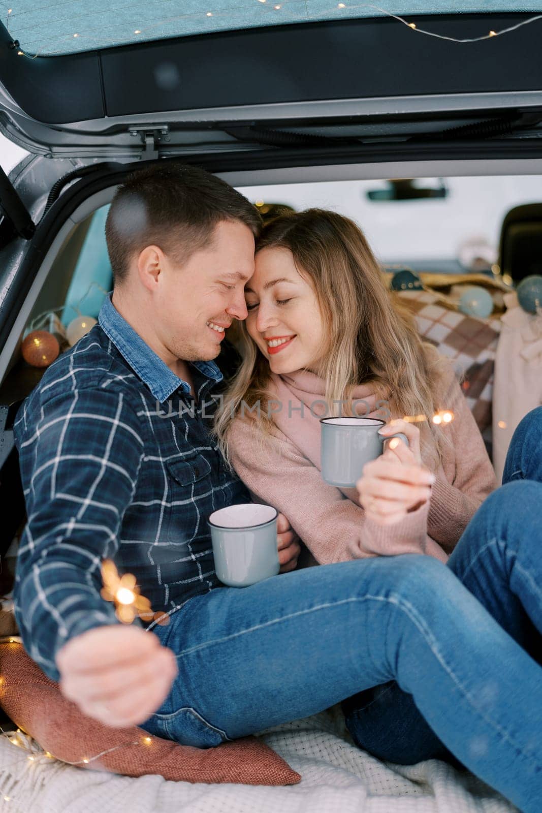 Smiling couple touching foreheads with mugs and sparklers while sitting in car trunk. High quality photo