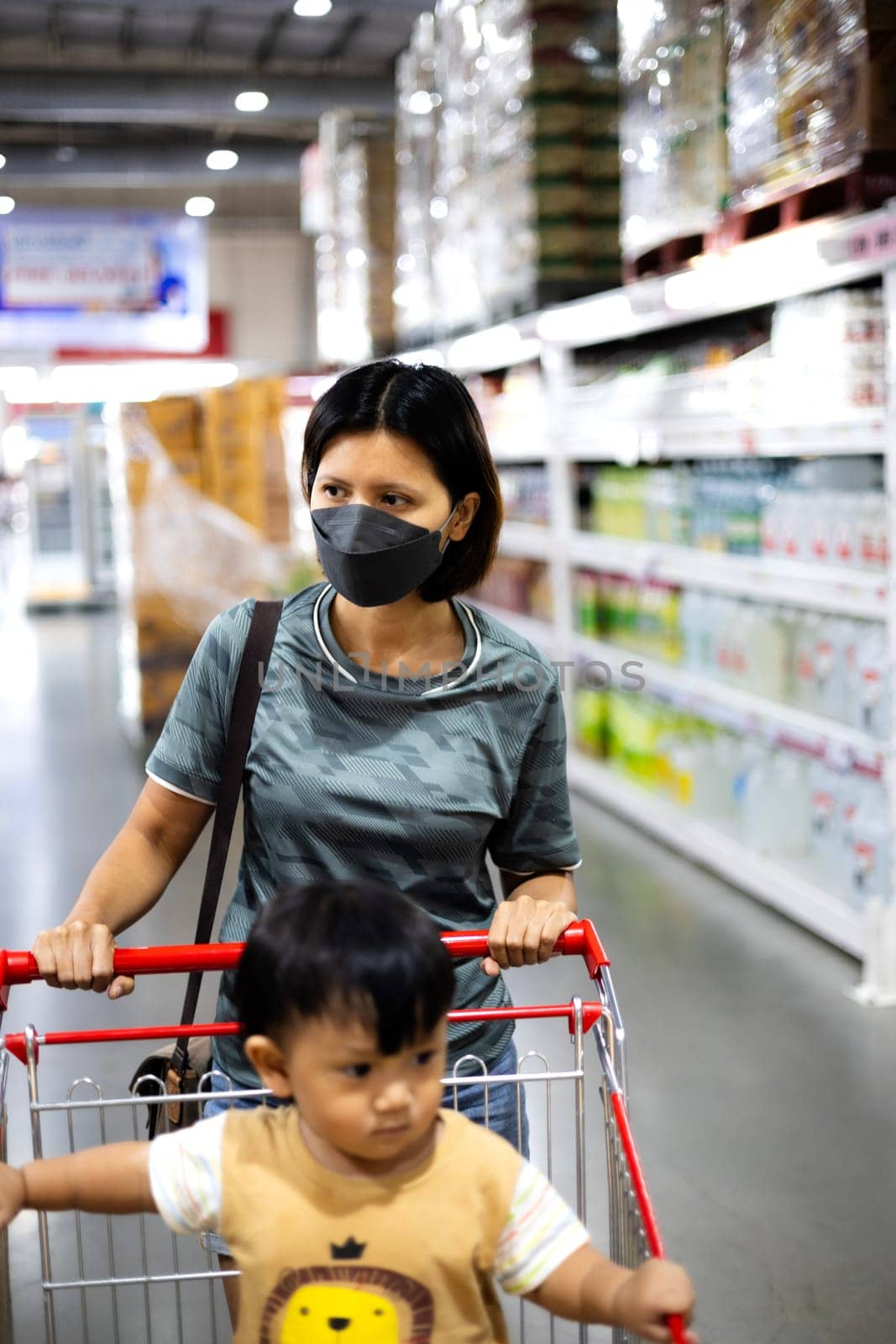 Mother pushing shopping cart with her infant baby boy child down department aisle in supermarket grocery store. Mother And Son At The Supermarket. Shopping with kids concept.