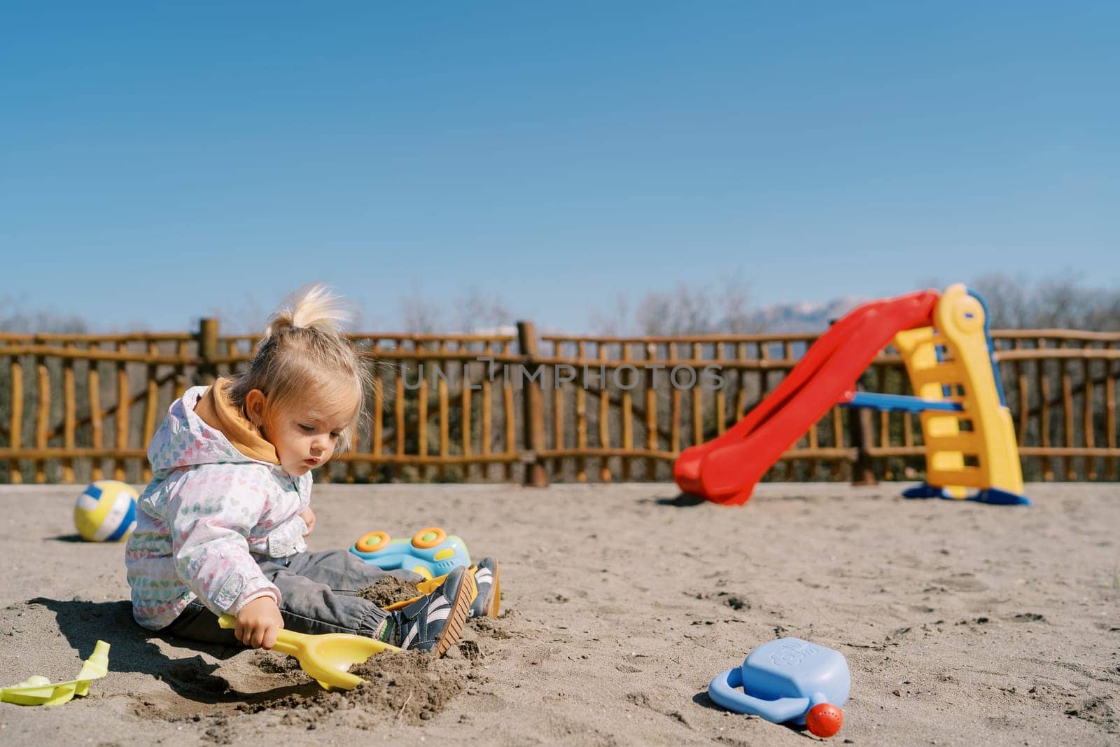 Little girl sits on the playground and digs the sand with a toy shovel by Nadtochiy