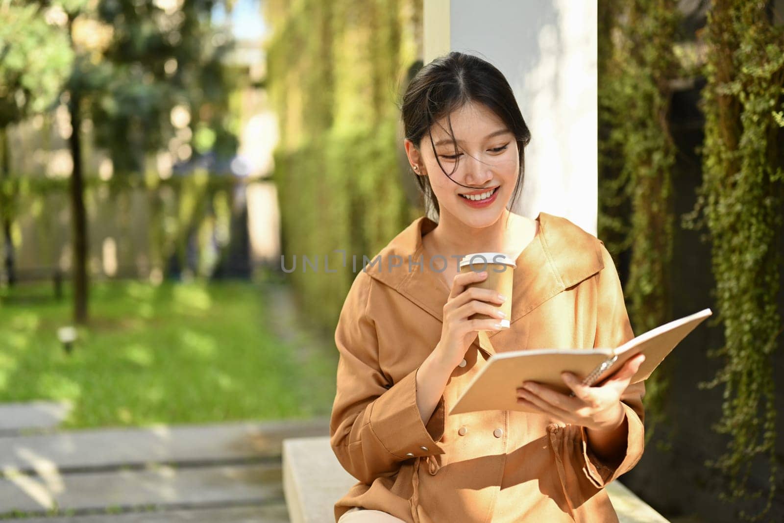 Pleased young Asian woman with take away coffee reading book outdoor on sunny day by prathanchorruangsak