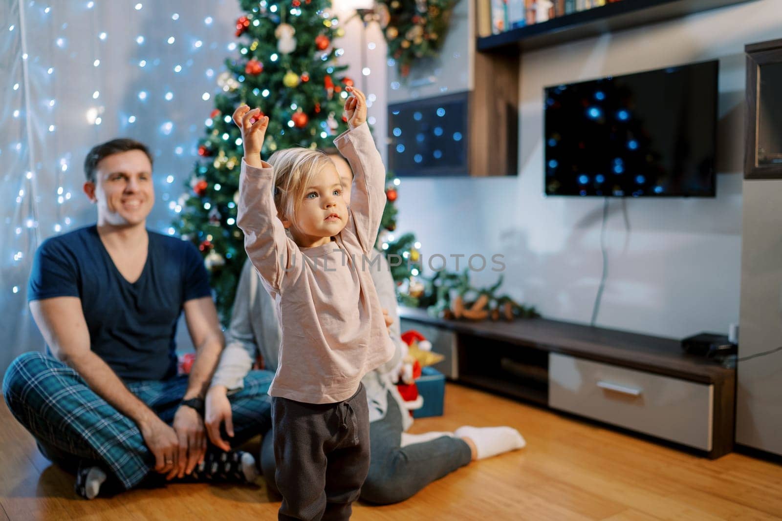 Little girl stands with raised hands near mom and dad sitting on the floor near the Christmas tree and looks away. High quality photo