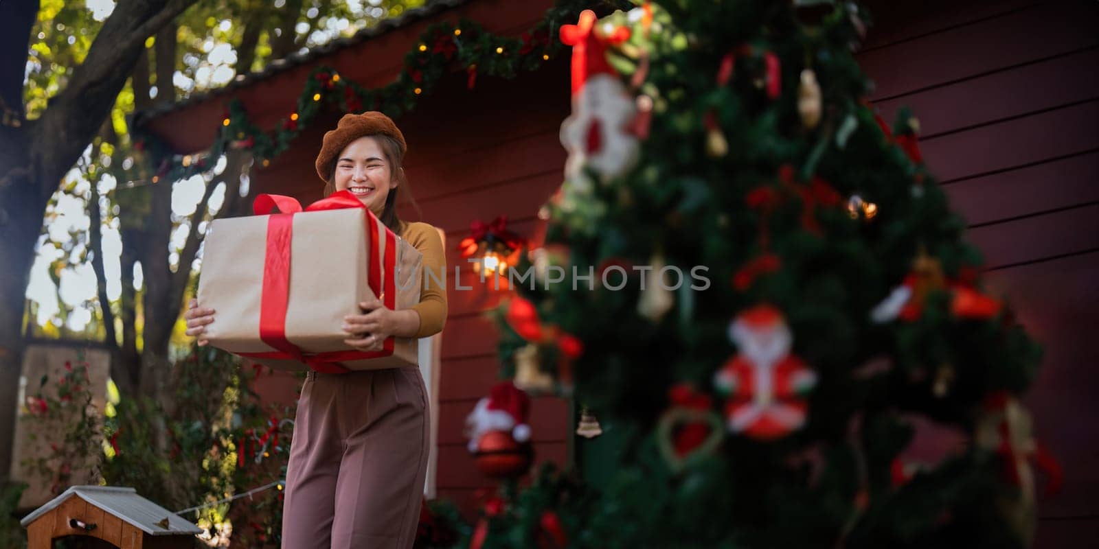 Happy woman wearing Santa hat holding of gift box. Positive emotional Santa girl. with a beautifully decorated Christmas tree serving as the background. festive Xmas concept by nateemee