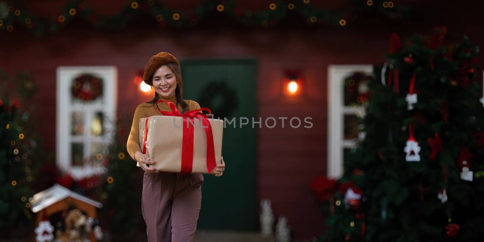 Happy woman wearing Santa hat holding of gift box. Positive emotional Santa girl. with a beautifully decorated Christmas tree serving as the background. festive Xmas concept.