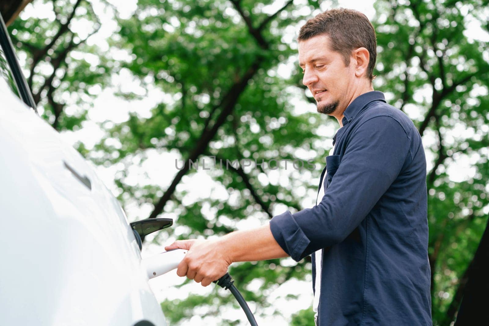 Man recharging battery for electric car during road trip travel EV car in natural forest or national park. Eco friendly travel during vacation and holiday. Exalt