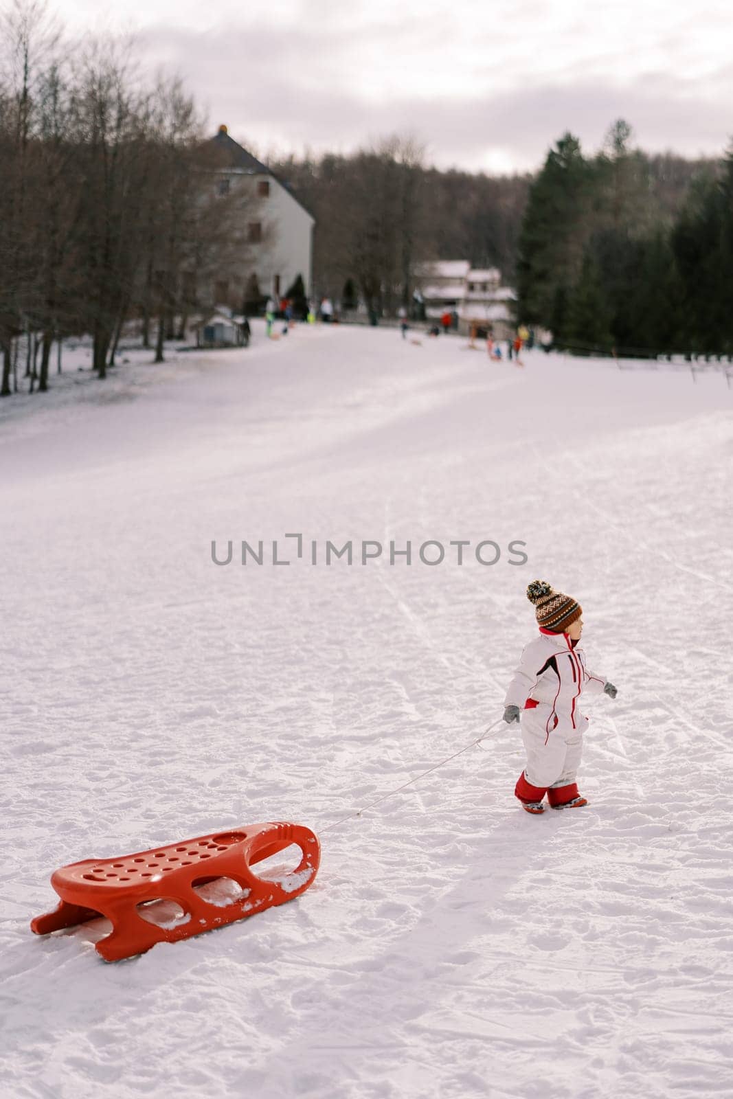 Little girl is walking along a snowy hill, dragging a sled on a rope behind her. Side view. High quality photo