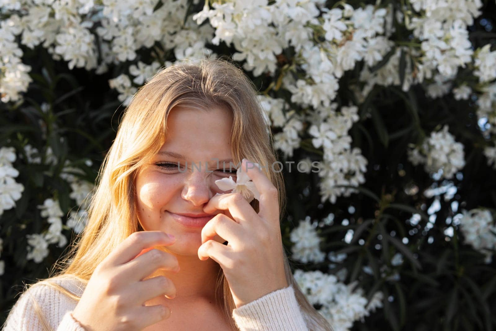 portrait of a teenage girl of European appearance, blond hair, the girl is standing on the street near a bush of white flowers, there is a place for an inscription. High quality photo