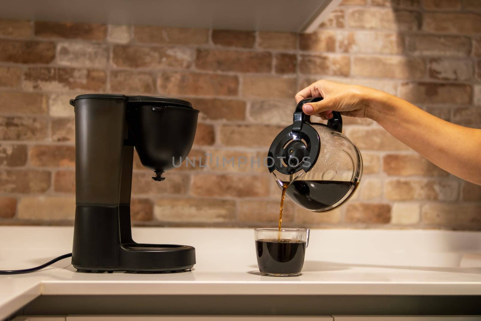 A woman's hand pours coffee from a coffee pot into a cup in the kitchen. Family life. Electric coffee maker on the kitchen countertop. There is a place for an inscription. High quality photo