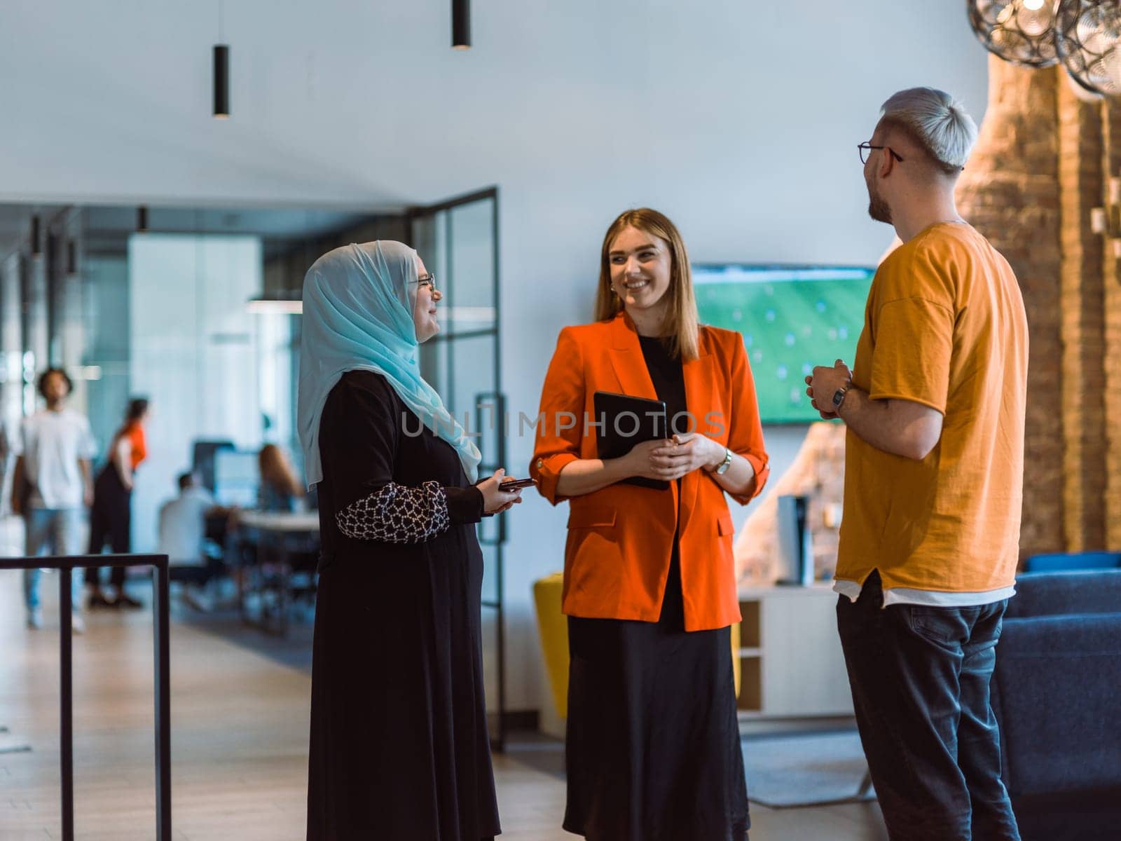 A group of young business colleagues, including a woman in a hijab, stands united in the modern corridor of a spacious startup coworking center, representing diversity and collaborative spirit by dotshock