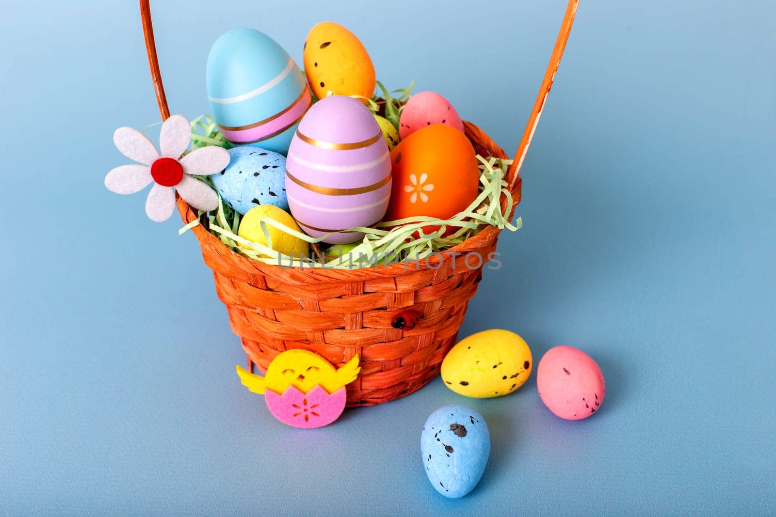 Close up of wicker basket filled with decorative multi colored easters eggs on a light blue background.