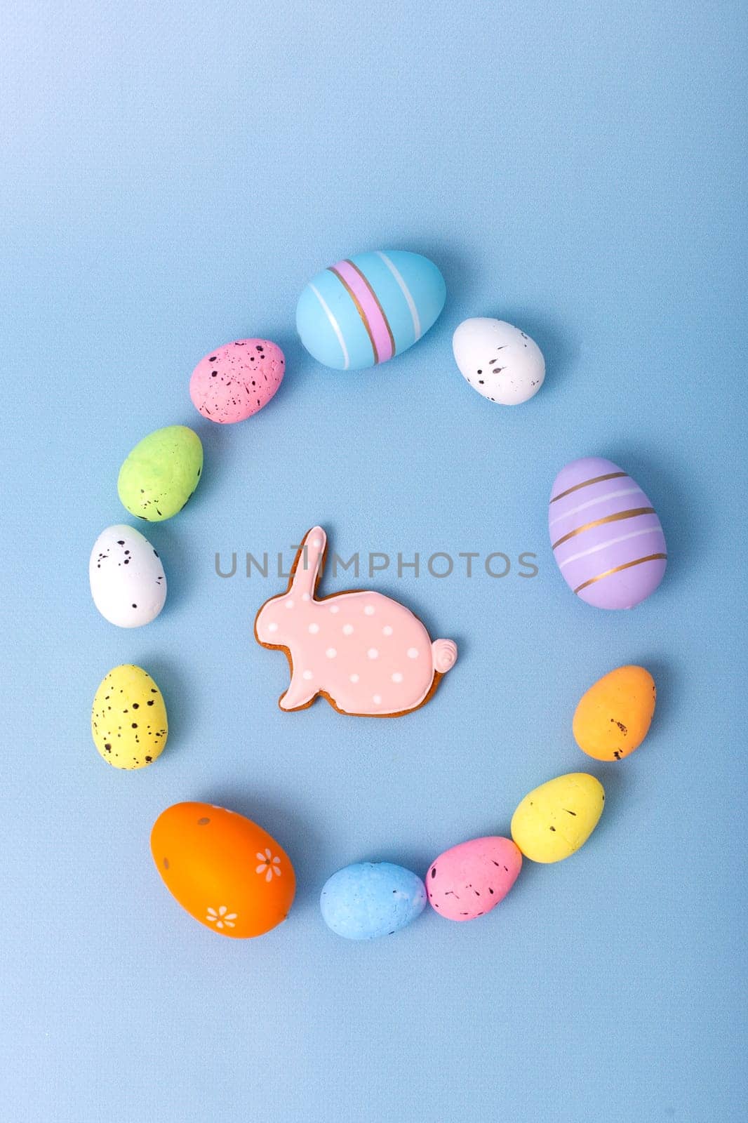 Beautiful decorative multicolored eggs and pink gingerbread in the form of a rabbit are scattered on a light blue background in an egg-shaped frame, top view