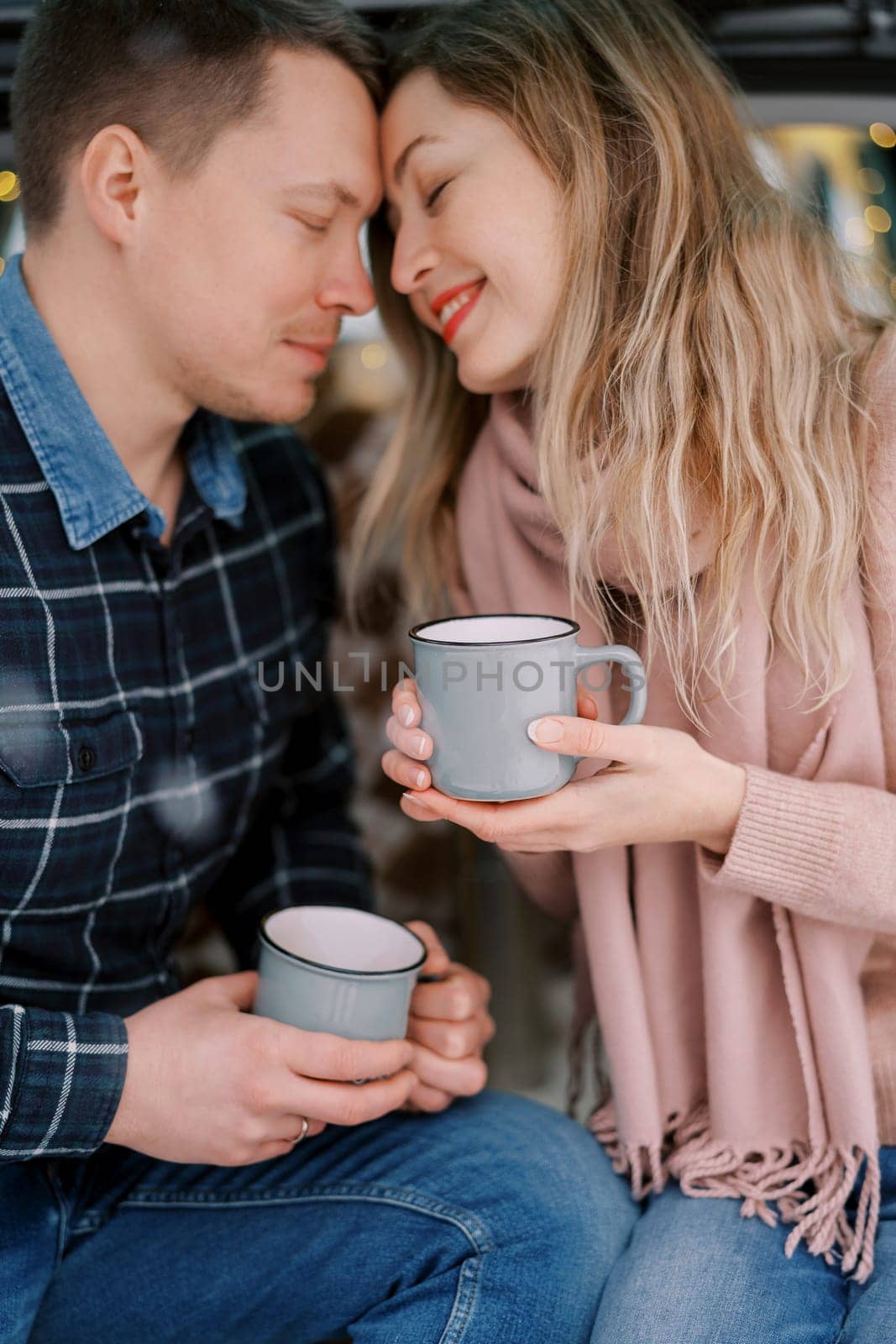 Smiling man and woman touching foreheads while sitting with cups in their hands by Nadtochiy