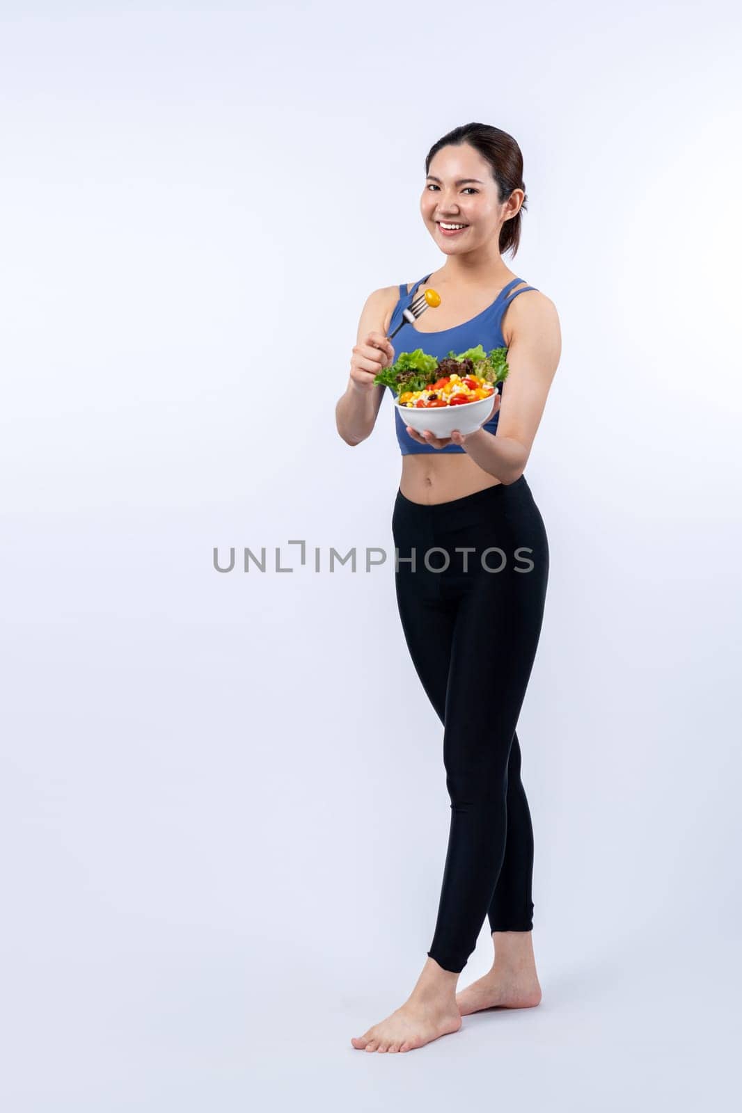 Young sporty Asian woman in sportswear holding salad bowl fill with vibrant of fruit and vegetable. Natural youthful and fit body lifestyle with balance nutrition on isolated background. Vigorous