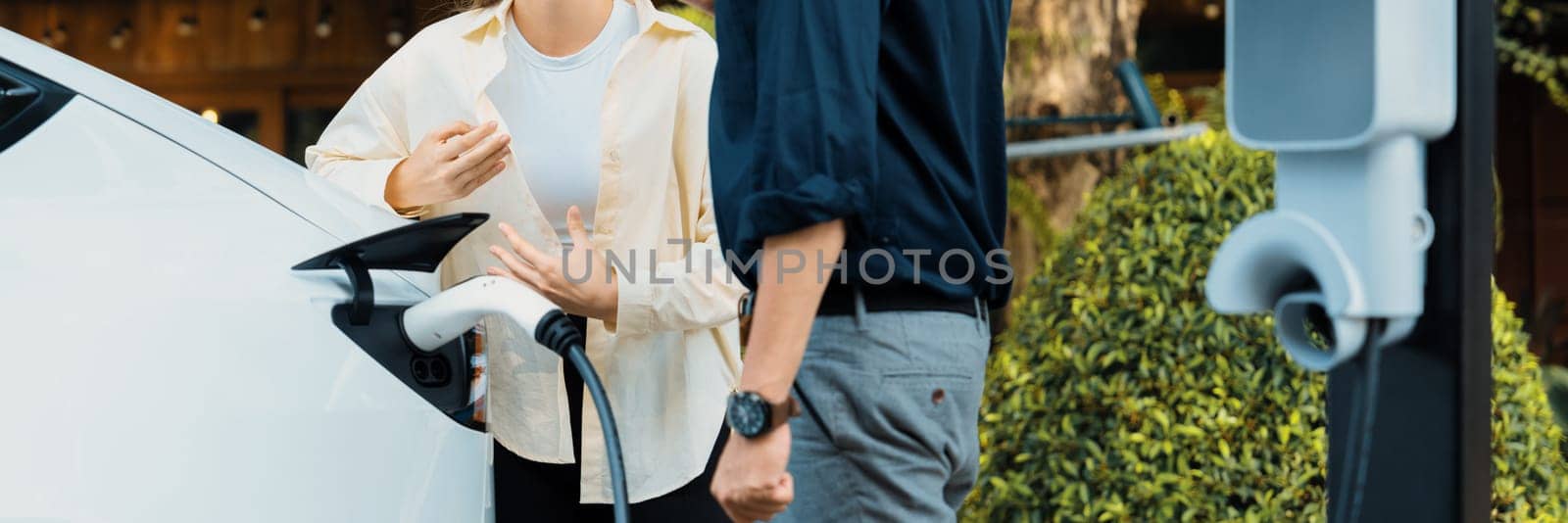 Young couple recharge electric car battery from charging station in green city park in springtime. Rechargeable EV car for sustainable environmental friendly urban travel lifestyle. Panorama Expedient