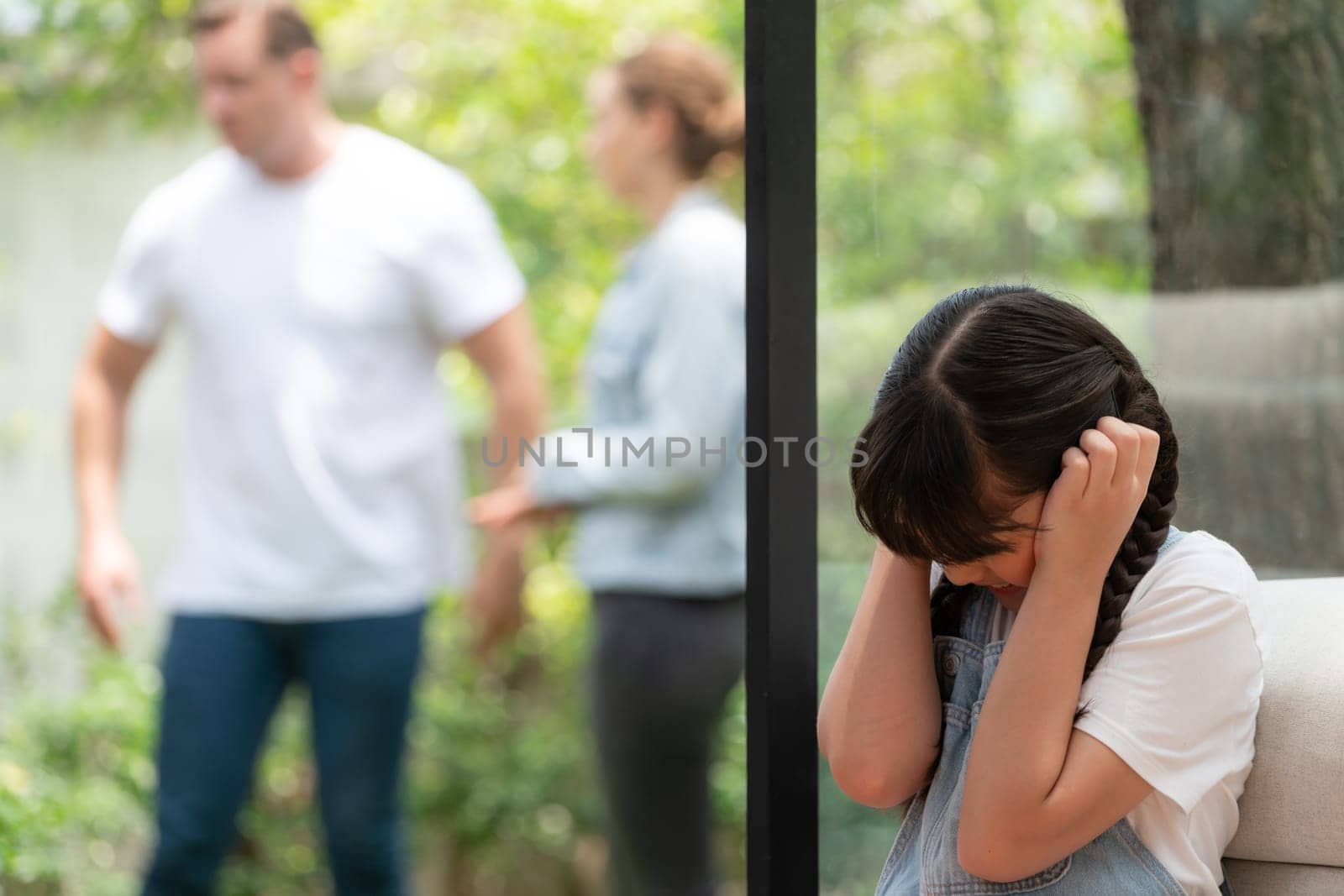 Stressed and unhappy young girl huddle in corner, cover her ears blocking sound of her parent arguing in background. Domestic violence at home and traumatic childhood develop to depression. Synchronos