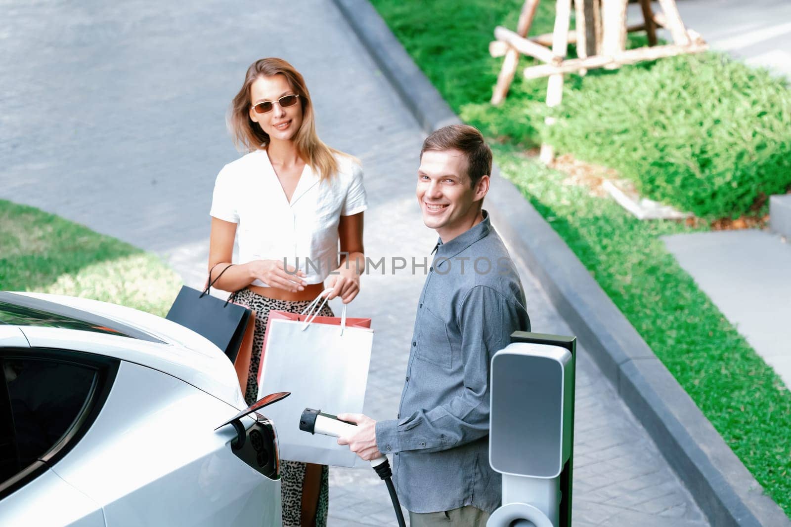 Young couple travel with EV electric car charging in green sustainable city outdoor garden in summer shows urban sustainability lifestyle by green clean rechargeable energy of electric vehicle innards