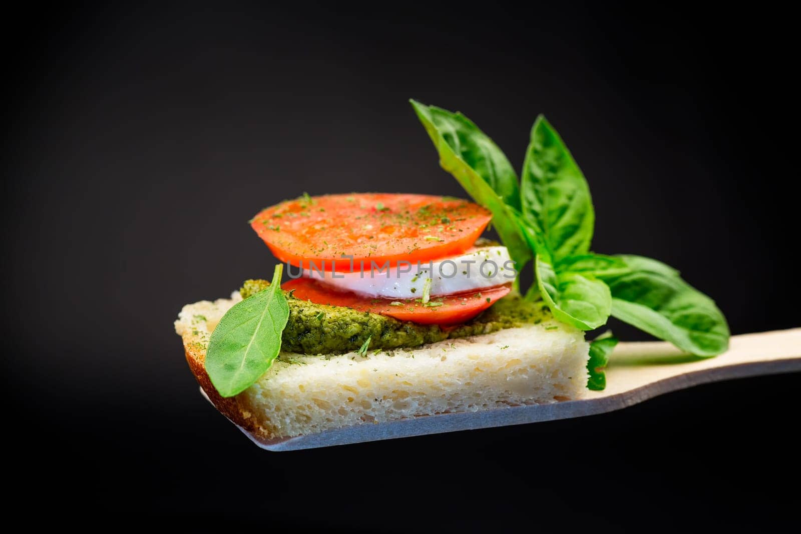 fresh slice of bread with pesto sauce, mozzarella and tomato on a wooden spatula with basil. Isolated on a black background.