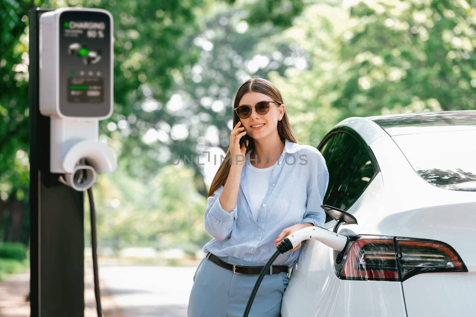 Young woman talking on smartphone while recharging electric car. Exalt by biancoblue